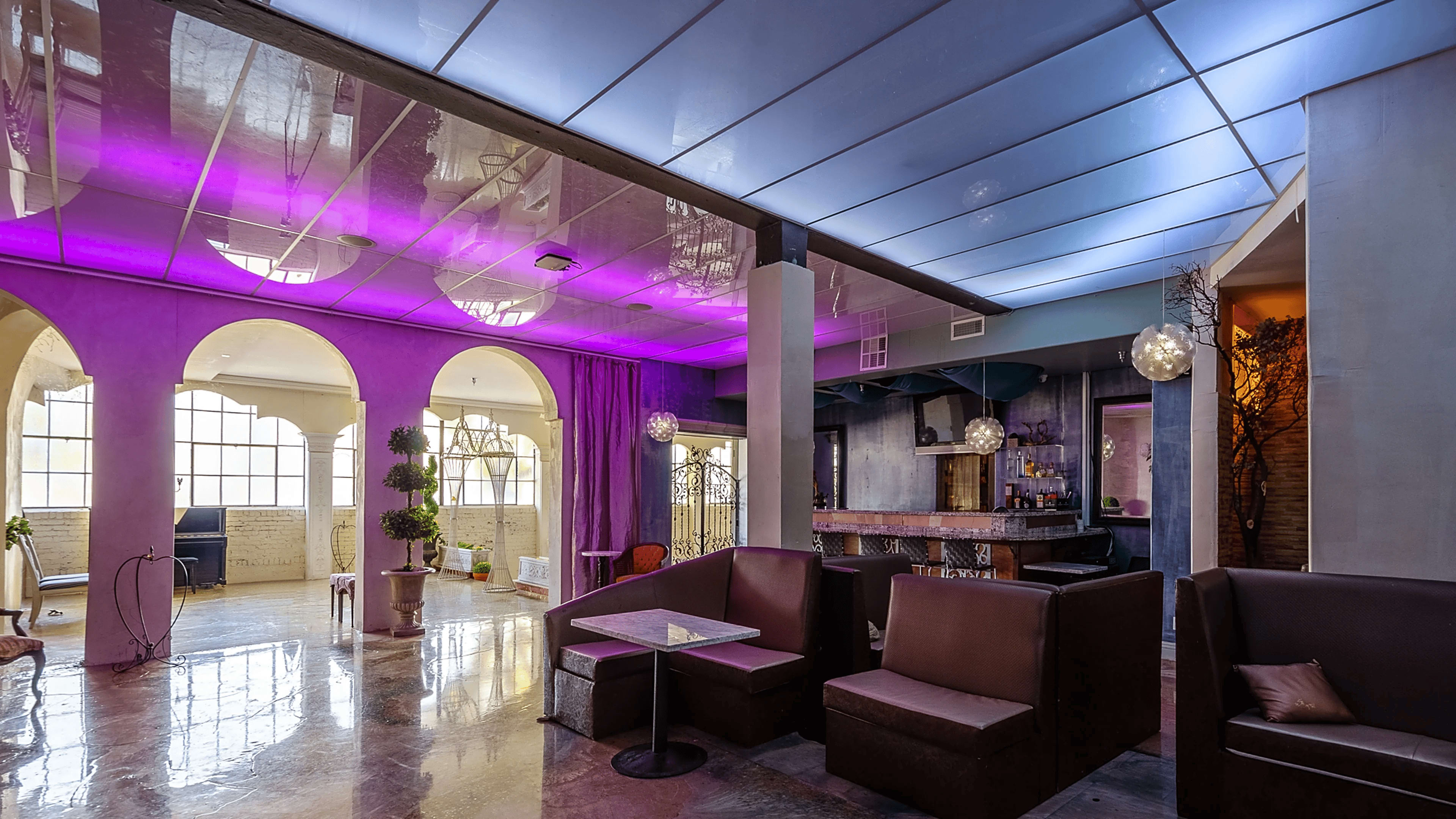 The image shows a modern lounge space featuring arched windows, a bar area, and seating arrangements with purple lighting accents.