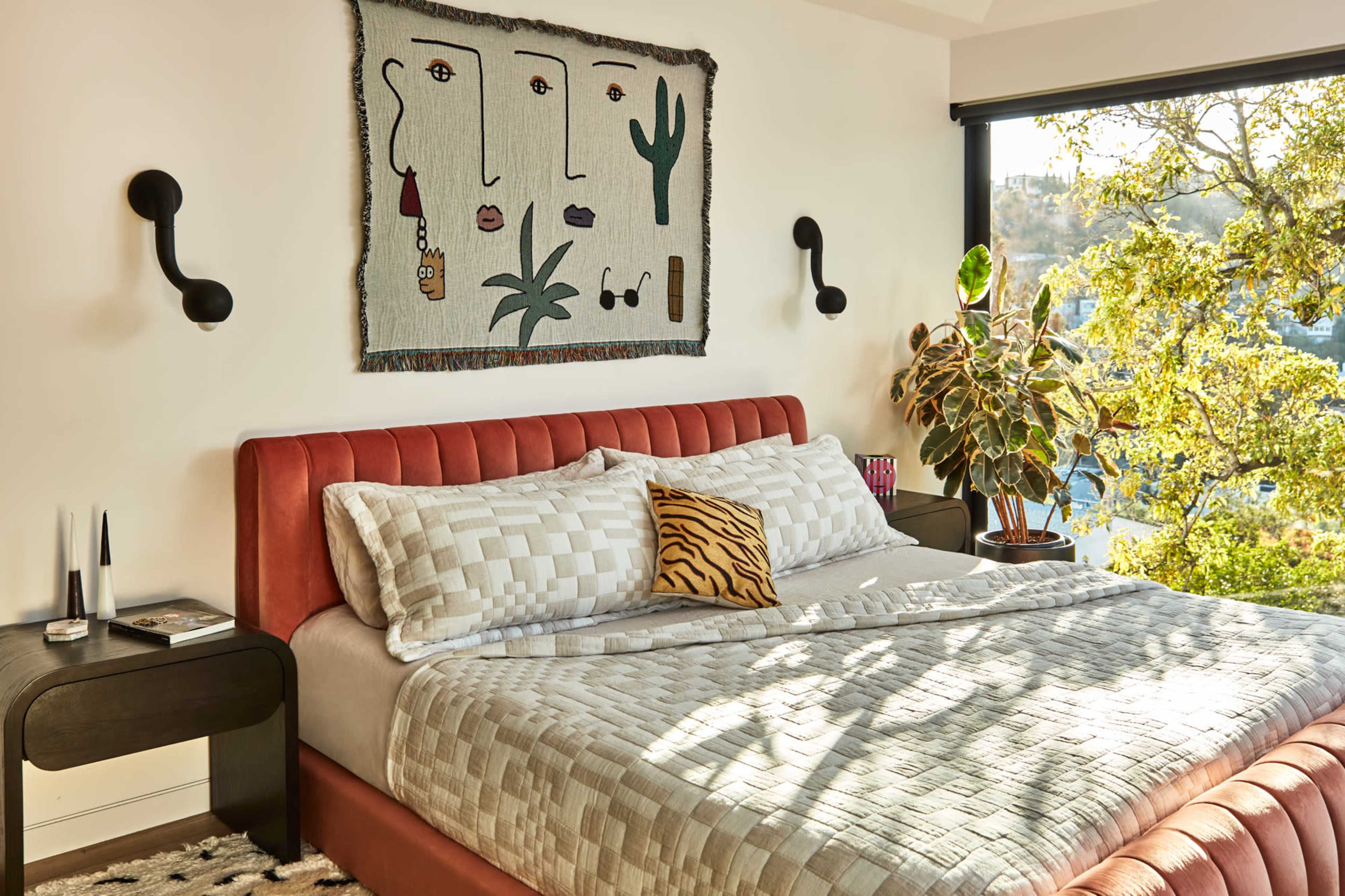 A modern bedroom features a pink upholstered bed with patterned bedding, a large wall tapestry, and a potted plant by a window overlooking greenery.