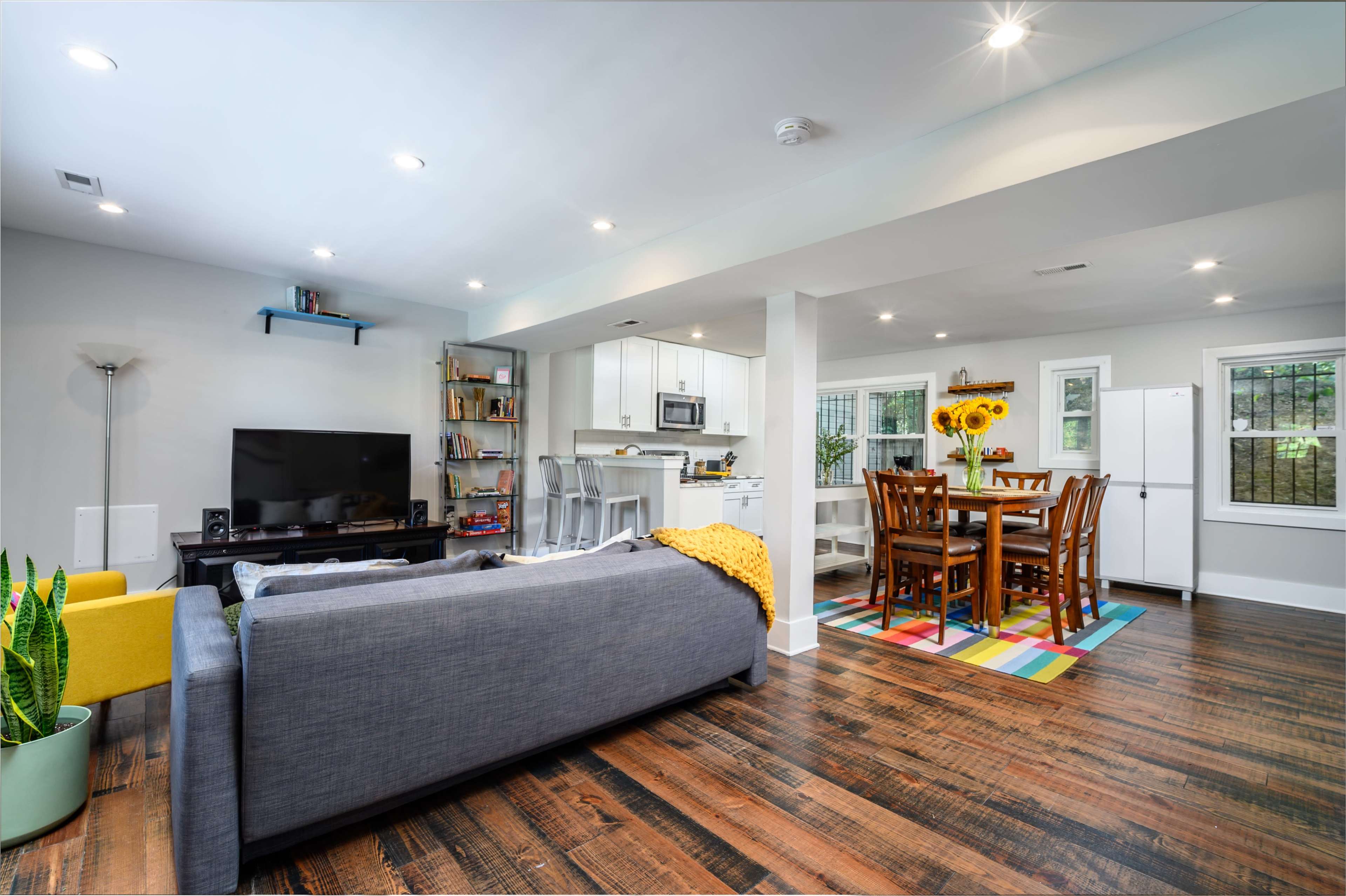 A modern living area features a gray sofa, television, and a dining space with a colorful rug, surrounded by bright windows and a kitchen area.