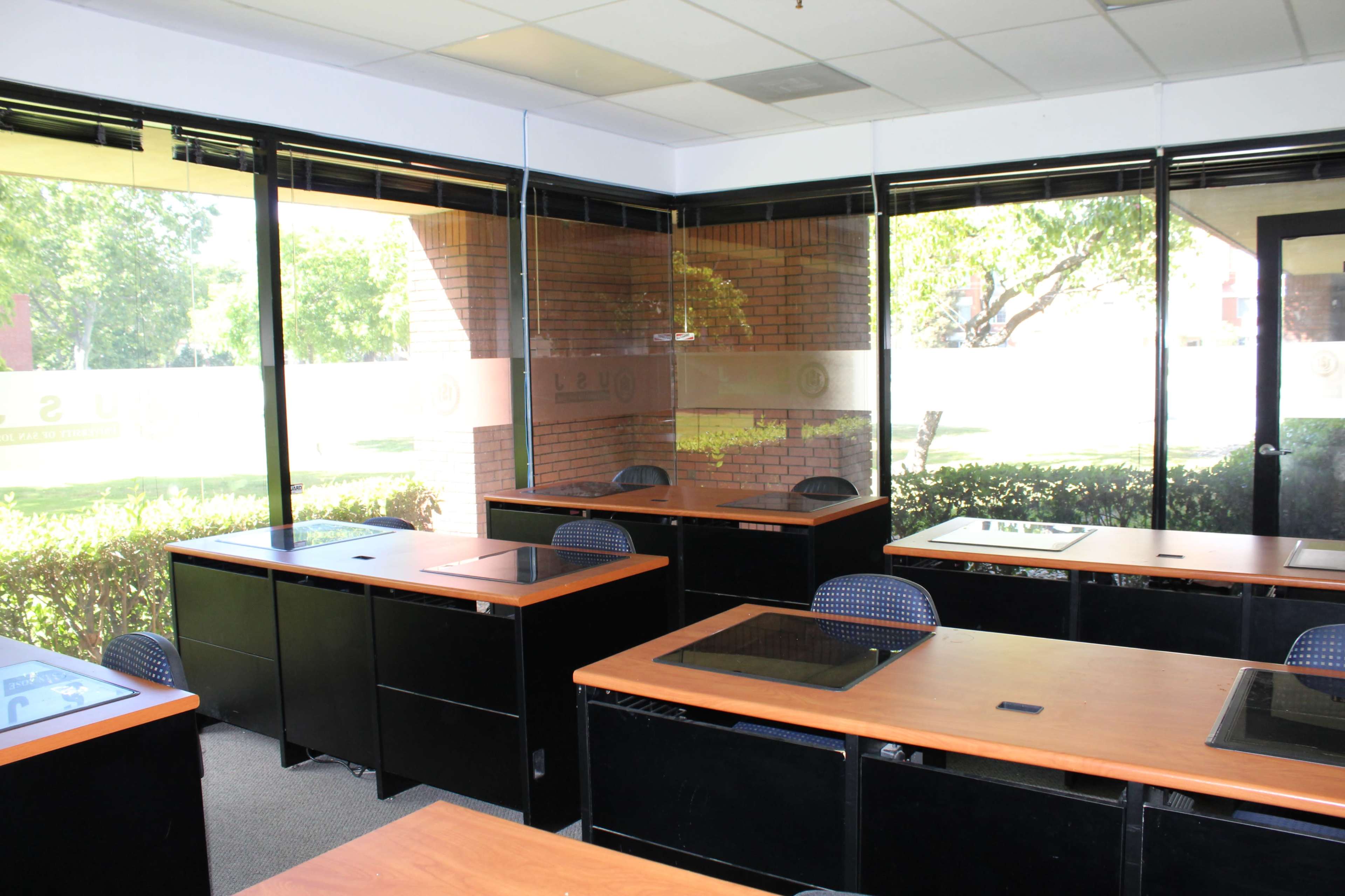 A modern classroom with several desks arranged in a well-lit space featuring large windows that overlook greenery.