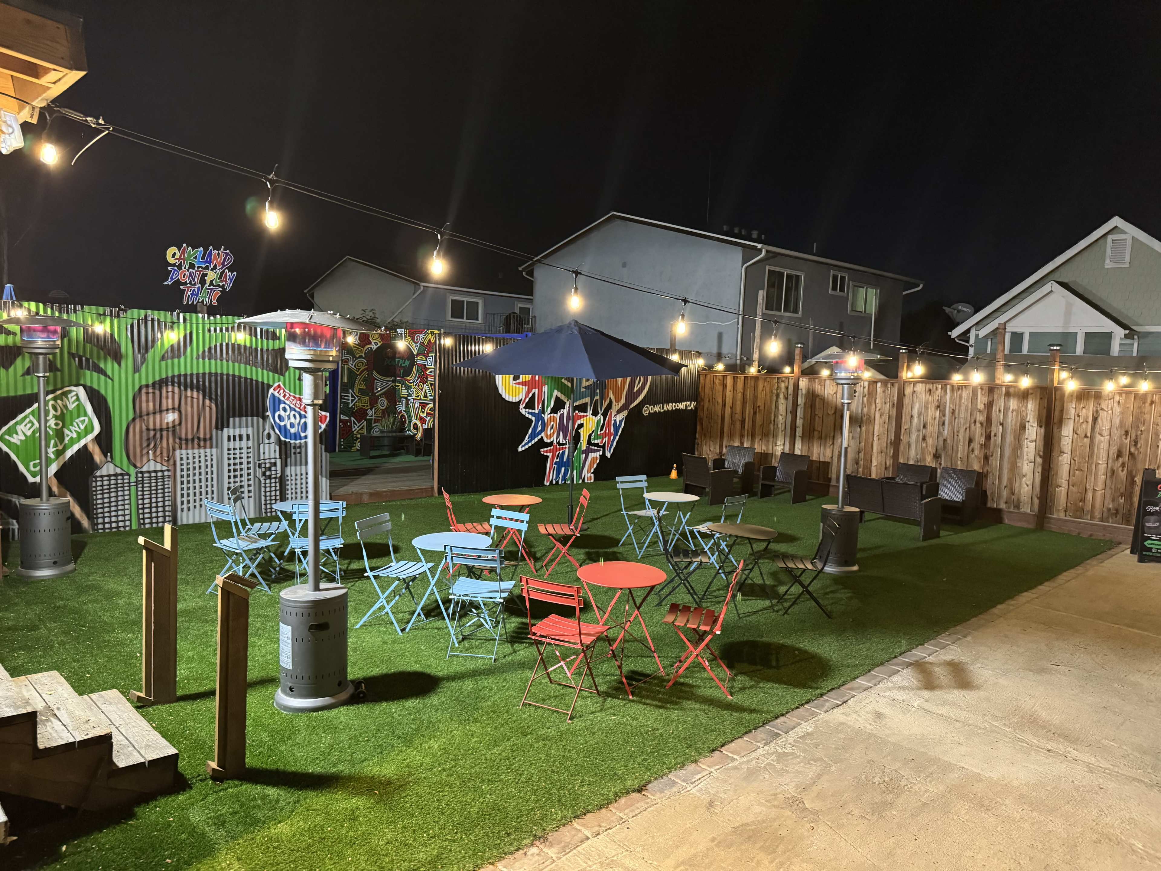 The image shows a backyard area at night with colorful tables and chairs, patio heaters, and string lights illuminating a vibrant mural on the wall.