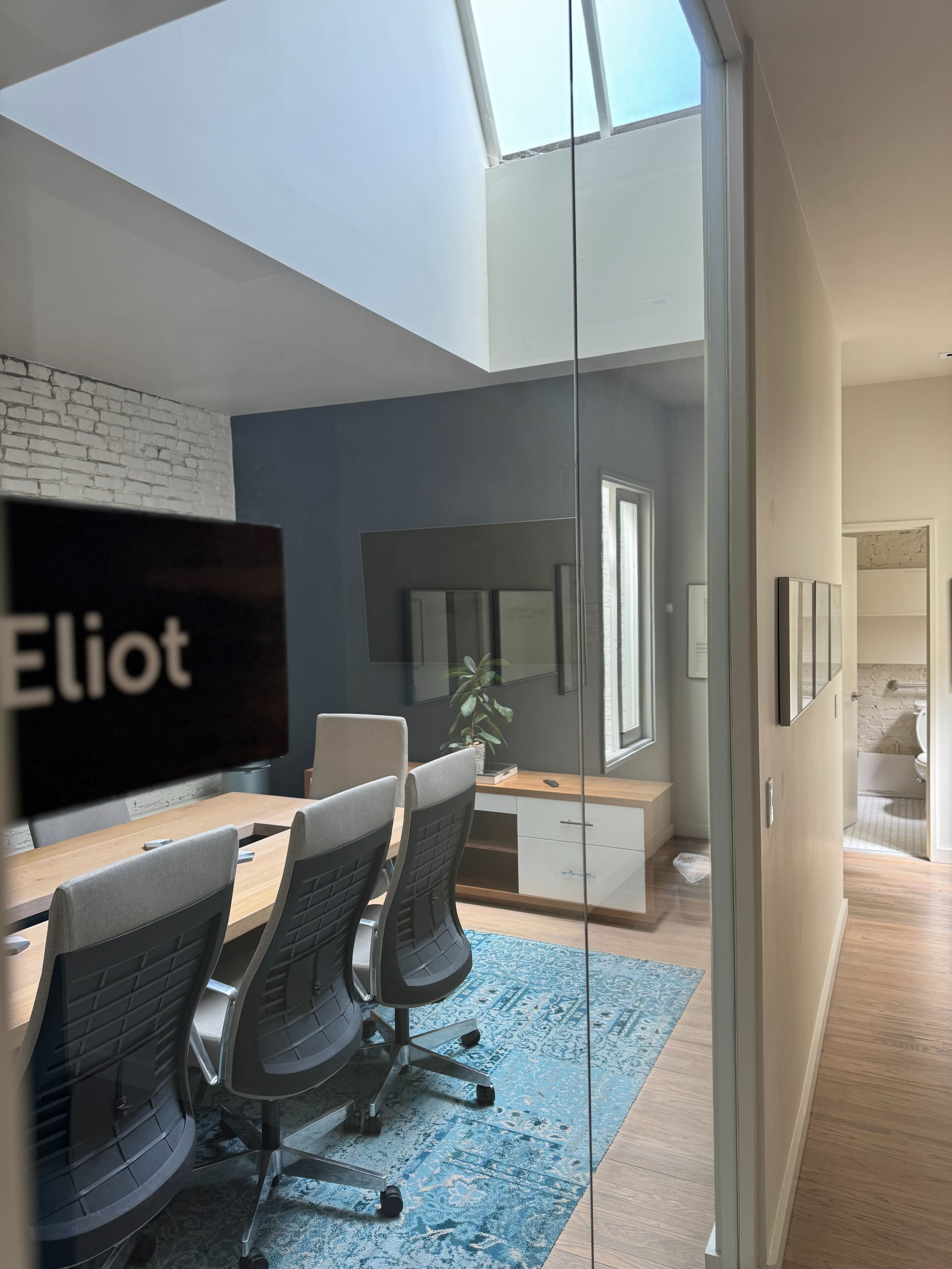 A modern conference room with a long table and ergonomic chairs, viewed through a glass wall.