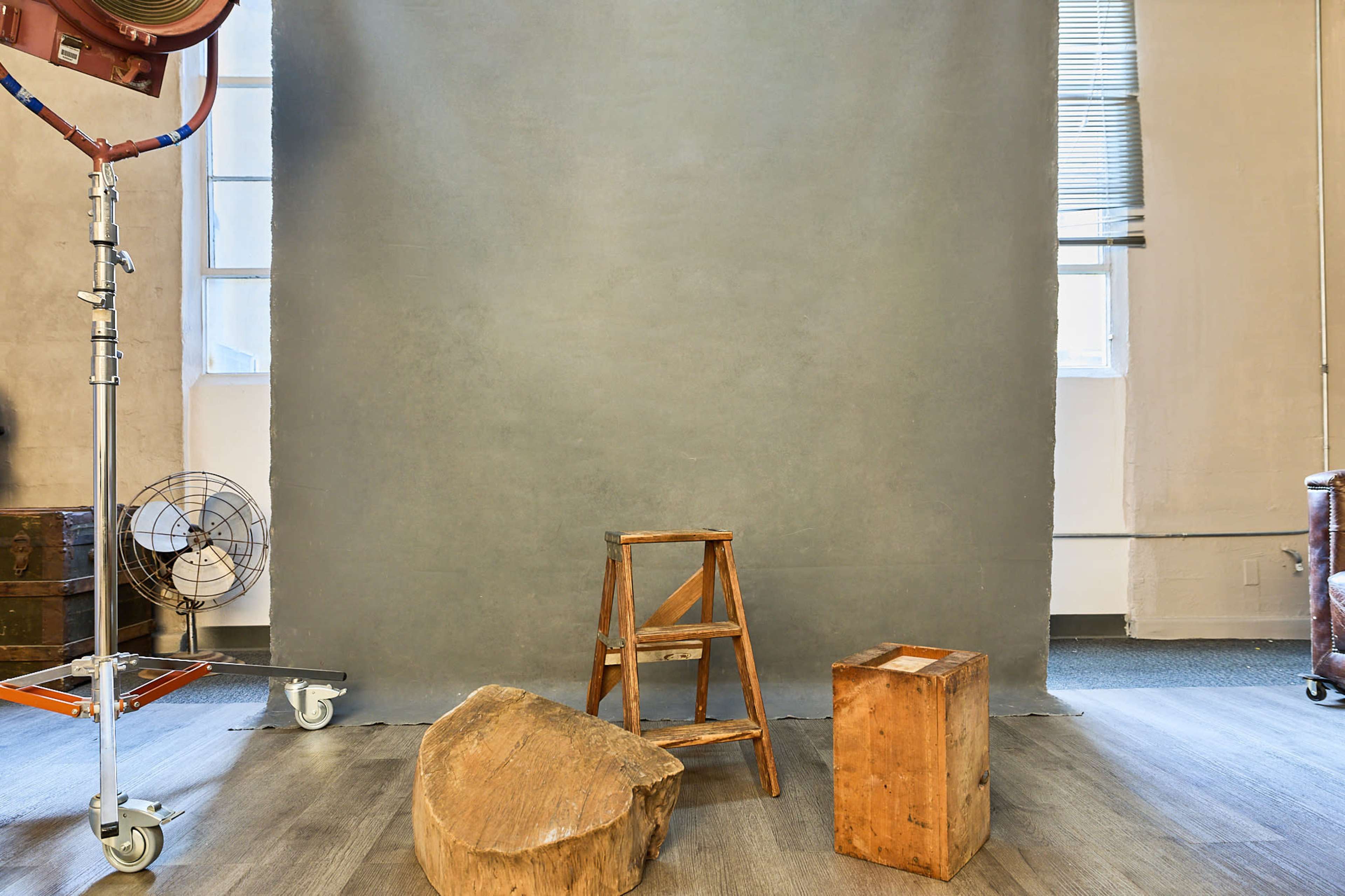 The image shows a photography studio with a gray backdrop, a wooden stool, a block of wood, and a floor fan.