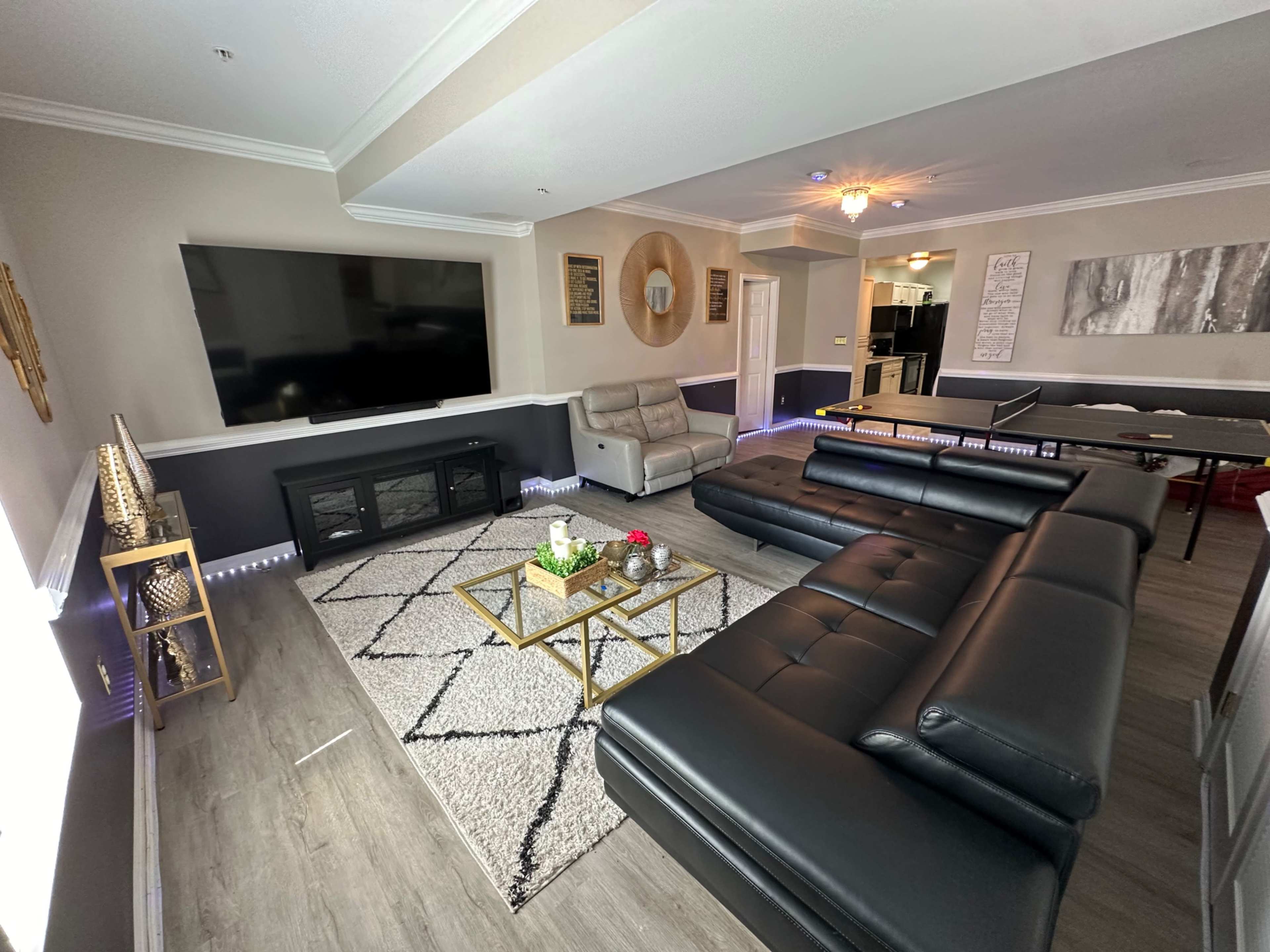 The scene features a modern living room with a large dark sectional sofa, a gold coffee table, a mounted flat-screen TV, and minimalistic decor against light-colored walls.