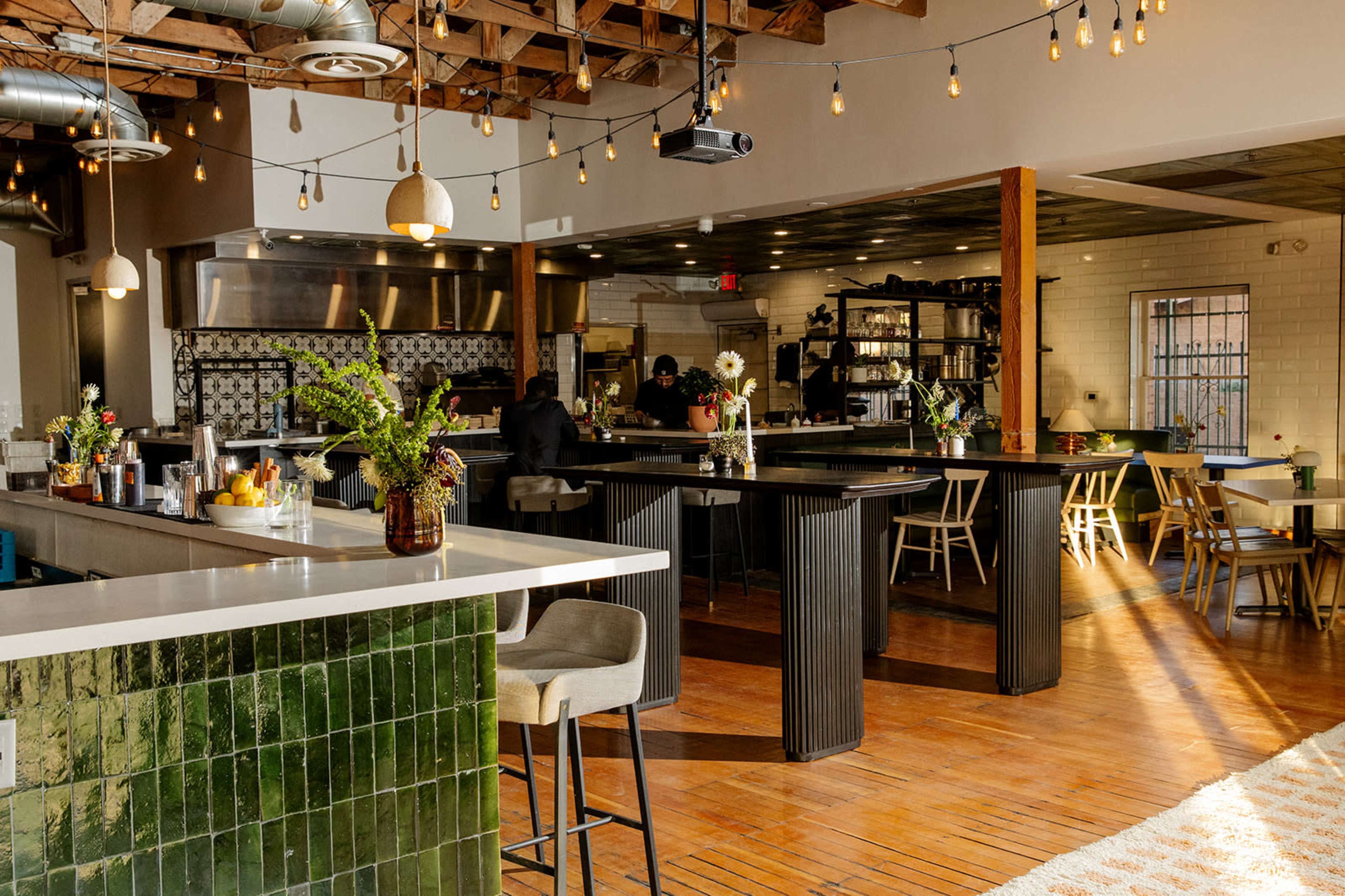 The image shows a modern restaurant interior with wooden floors, a decorative bar area, and pendant lighting, featuring tables and chairs arranged for dining.