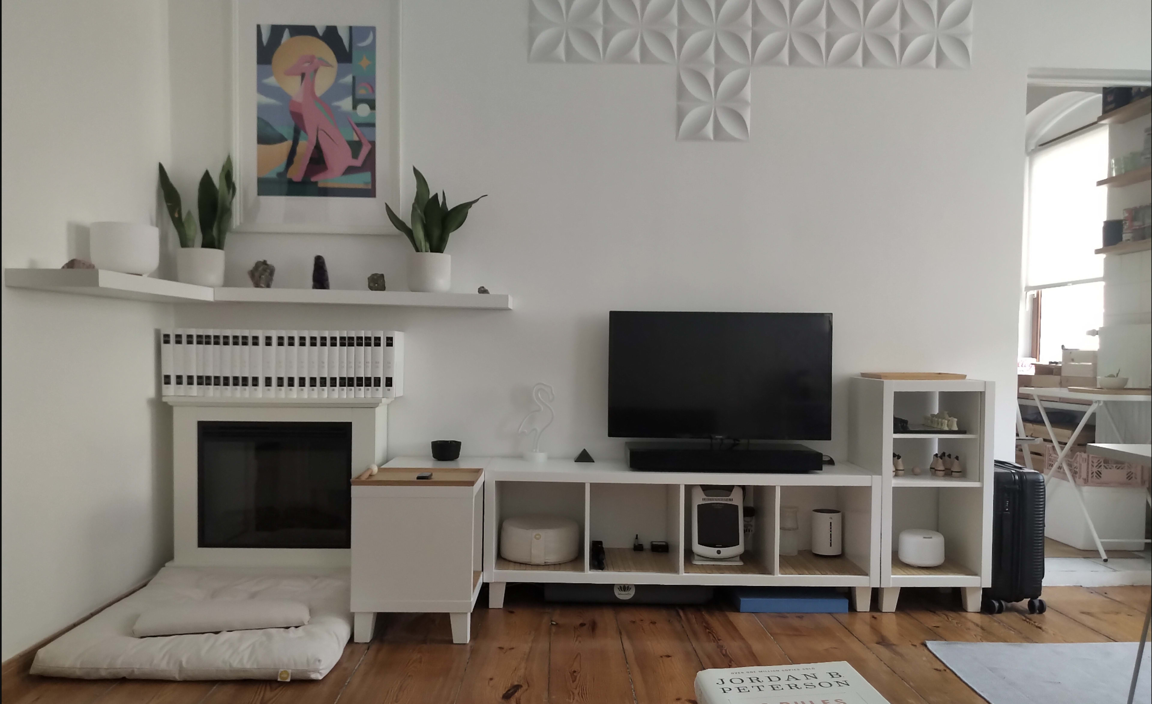 The image shows a living room with a white wall featuring decorative panels, a television on a white media console, a fireplace, and a framed artwork above the mantel.