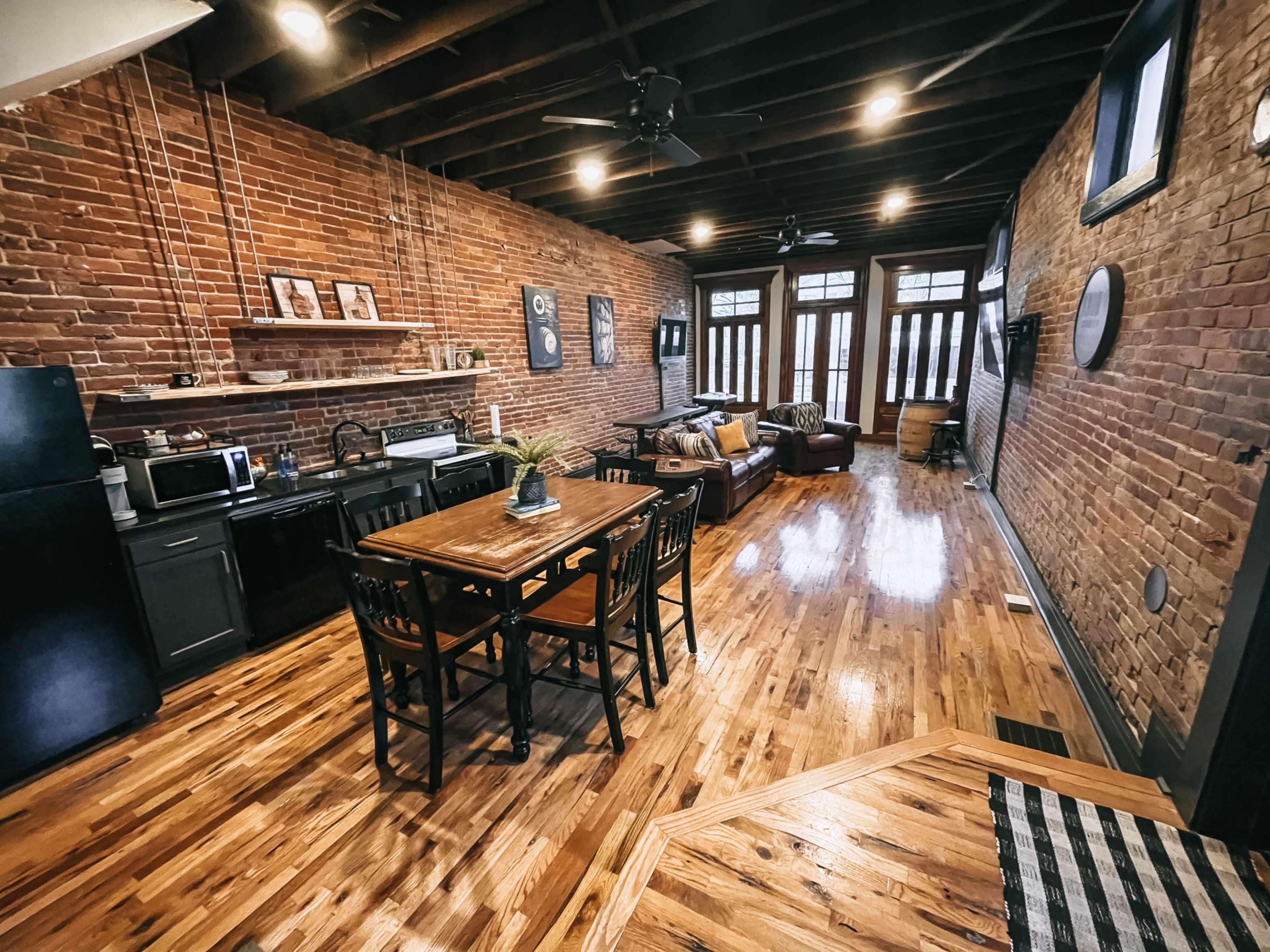 A spacious, open-concept living area featuring exposed brick walls, a wooden dining table with chairs, a kitchen area, and large windows at the far end.