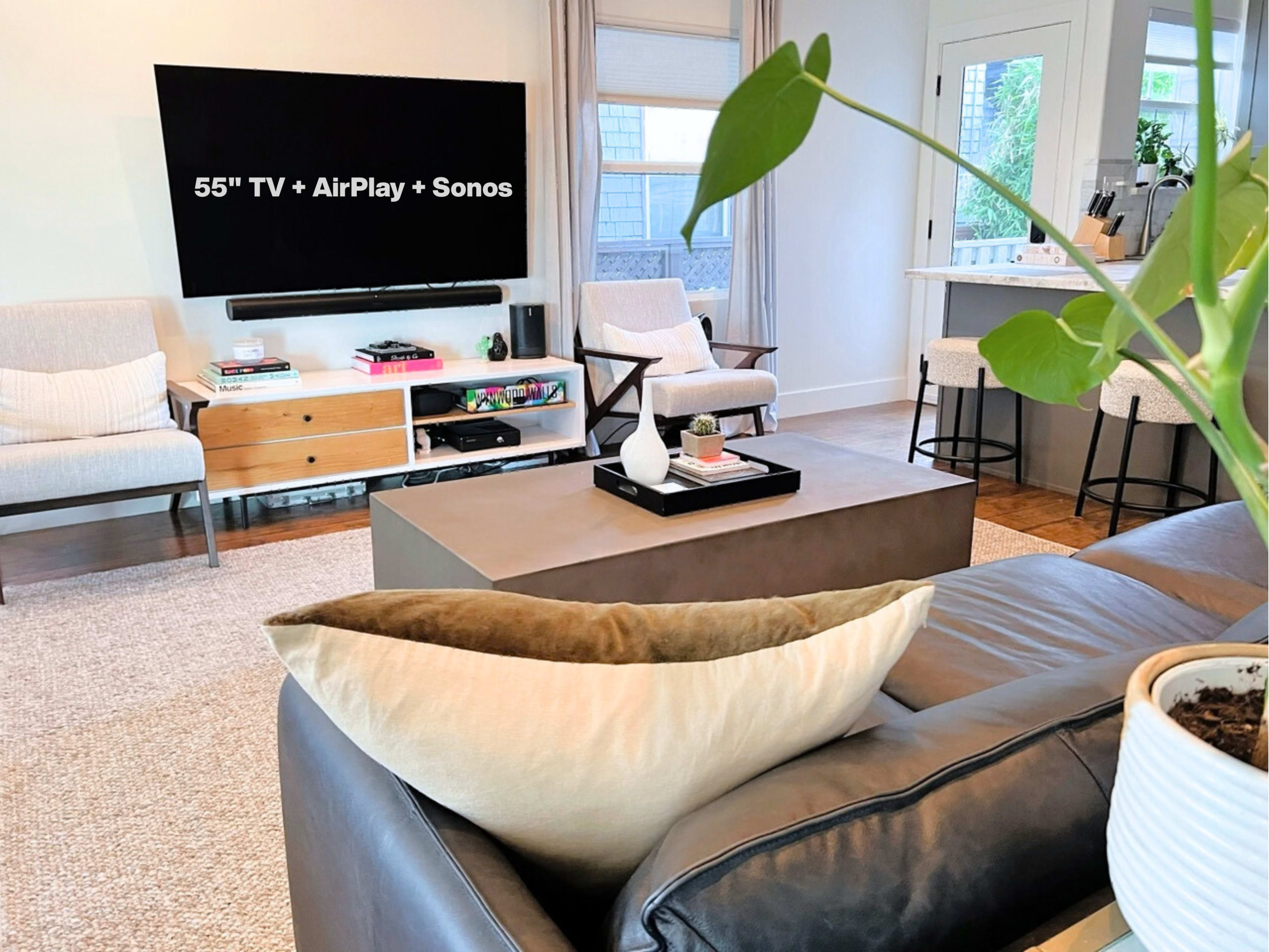 The image shows a modern living room featuring a 55-inch TV mounted on the wall, a coffee table in the center, and two armchairs beside a kitchen bar counter.