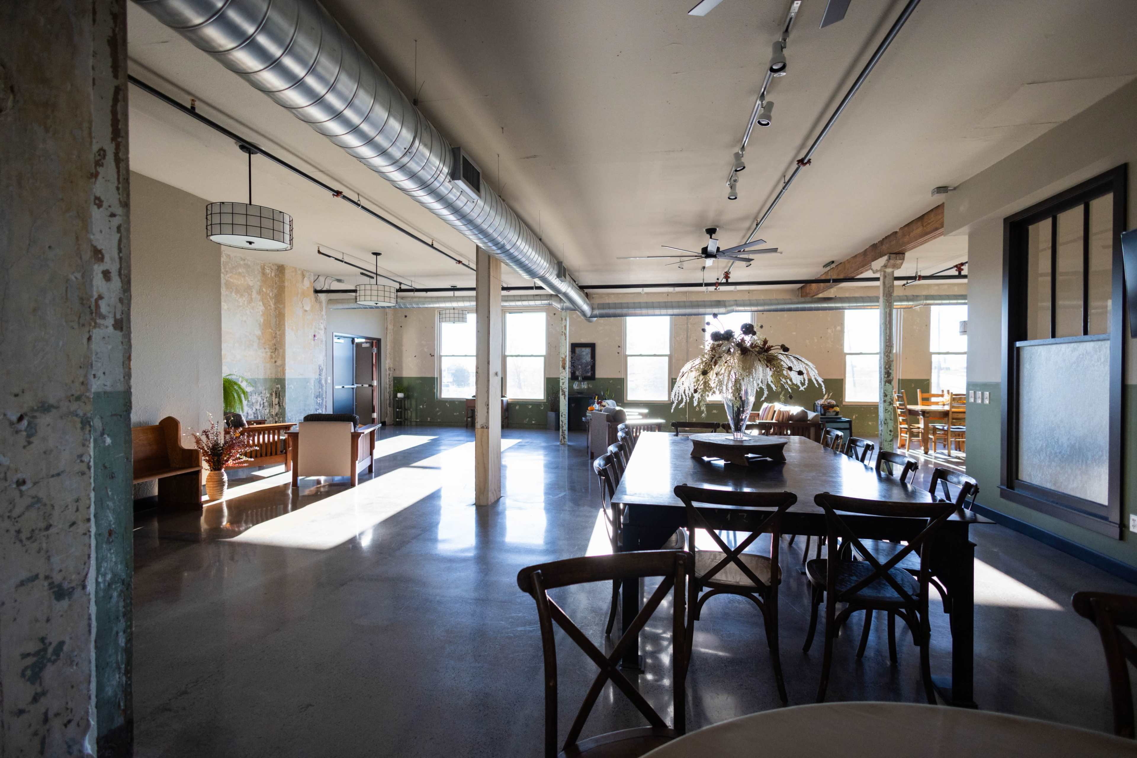 An industrial-style interior with large windows, a long dining table, and a mix of seating areas.