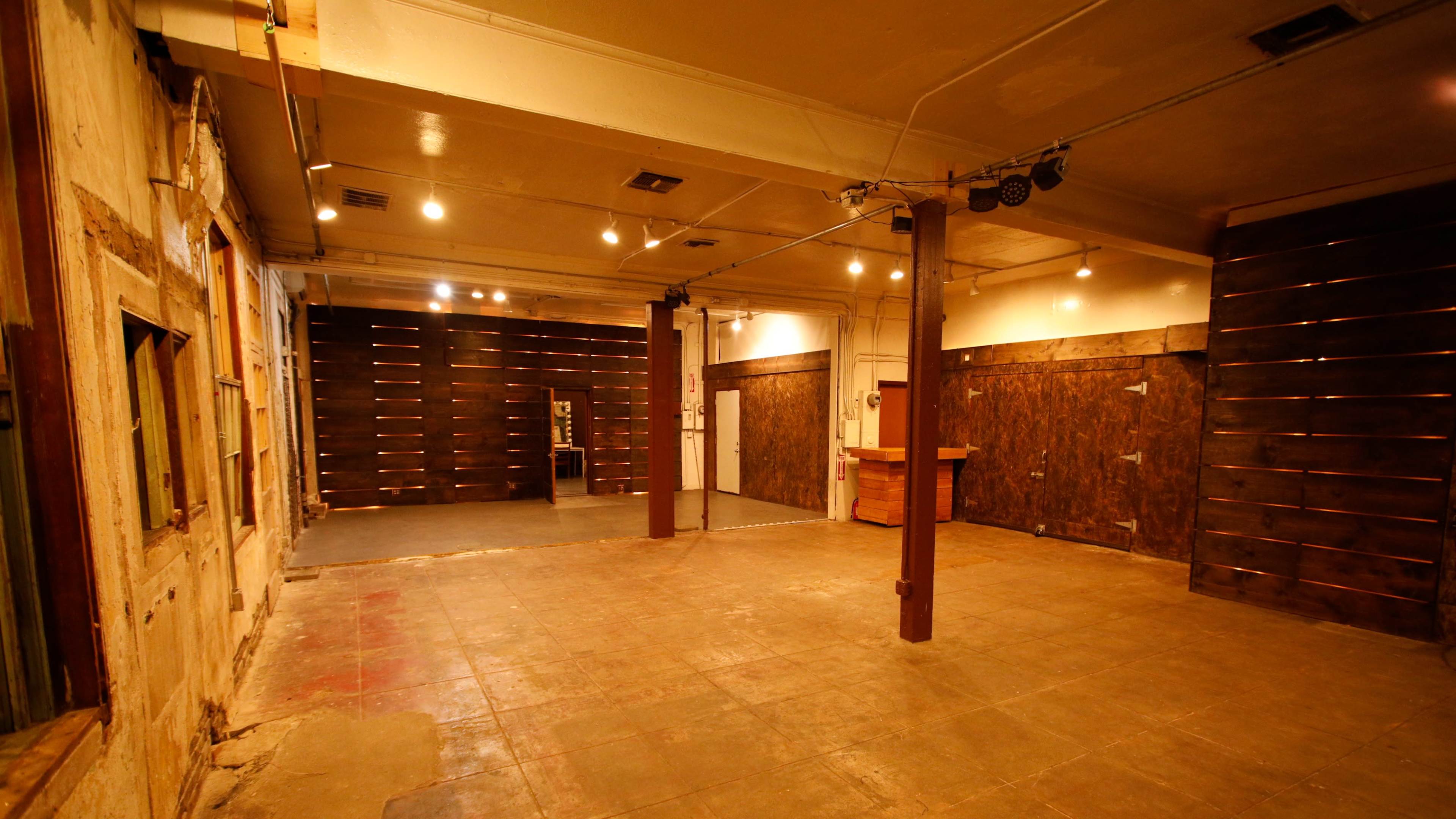 A spacious, empty interior with wooden walls and a concrete floor, featuring overhead lighting and a bar area in the corner.