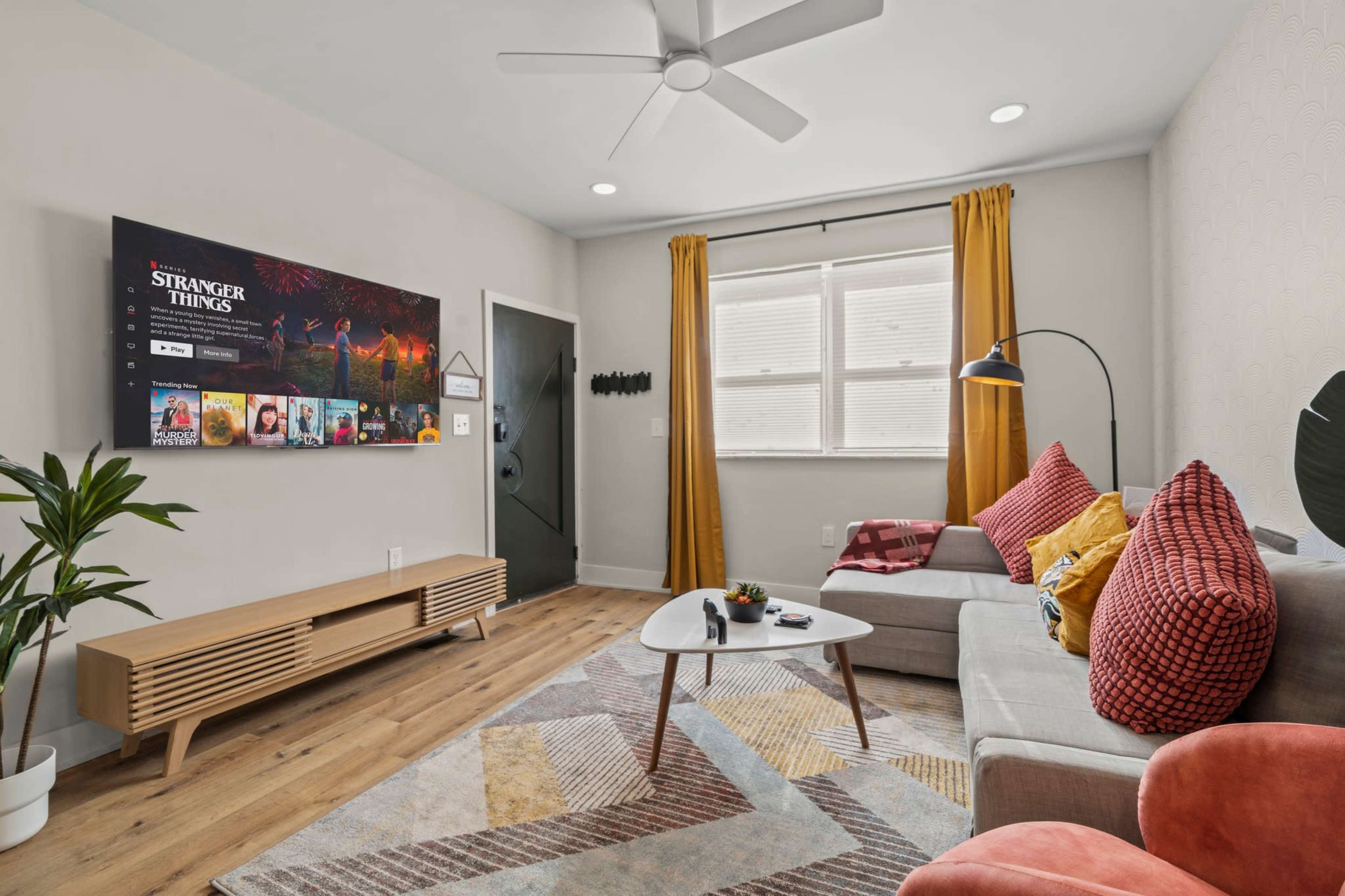 A modern living room features a sectional couch, a coffee table with remote controls, a large TV mounted on the wall displaying a streaming service, and bright yellow curtains framing a window.