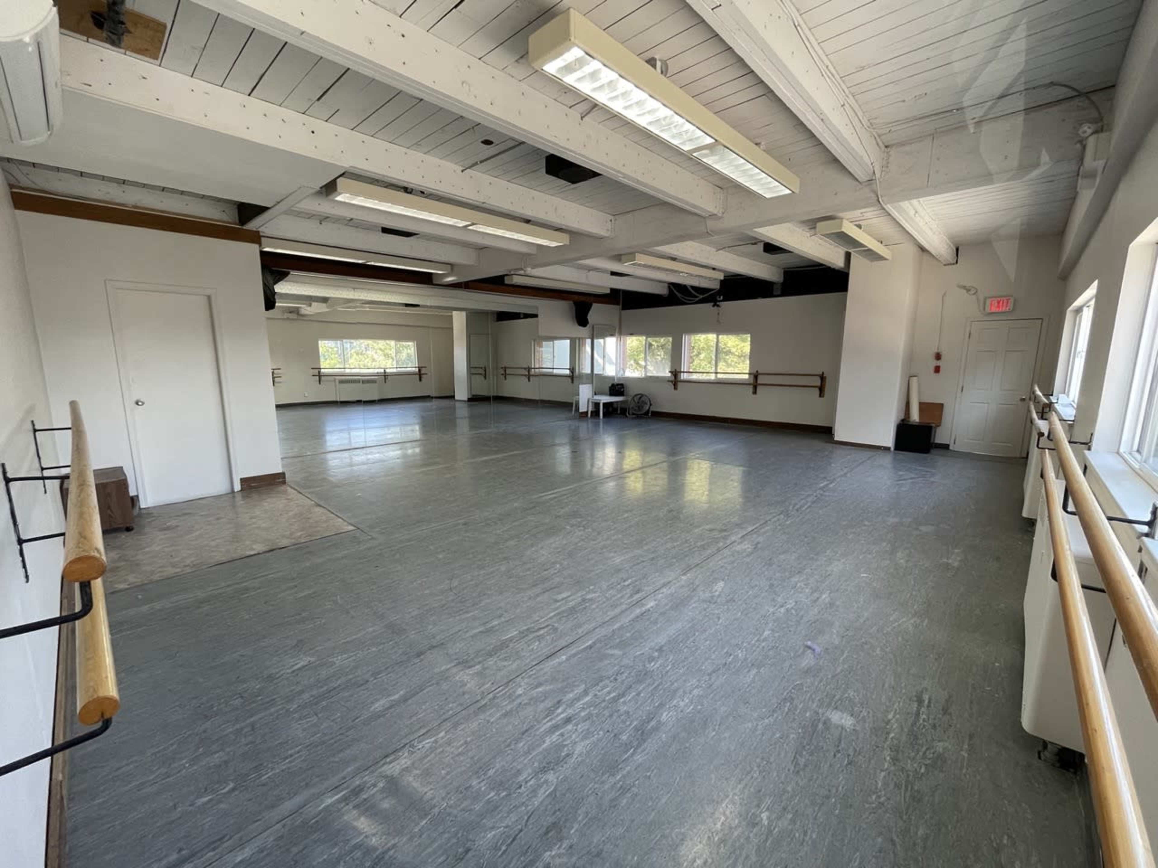 The image shows a spacious, well-lit dance studio with grey flooring, large windows, and mirrored walls.