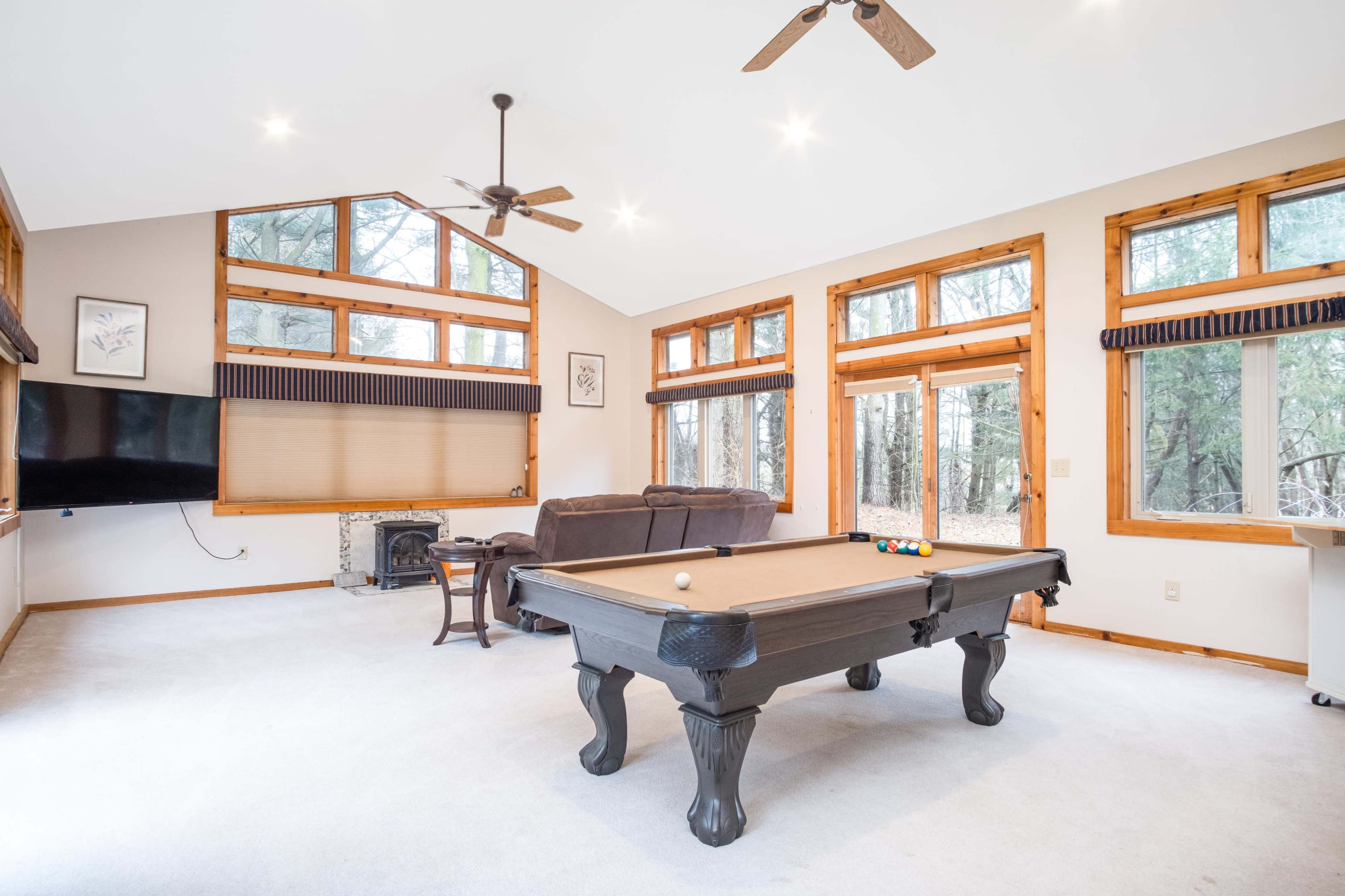 The spacious room features a pool table, large windows, and a flat-screen TV, with views of the surrounding forest.