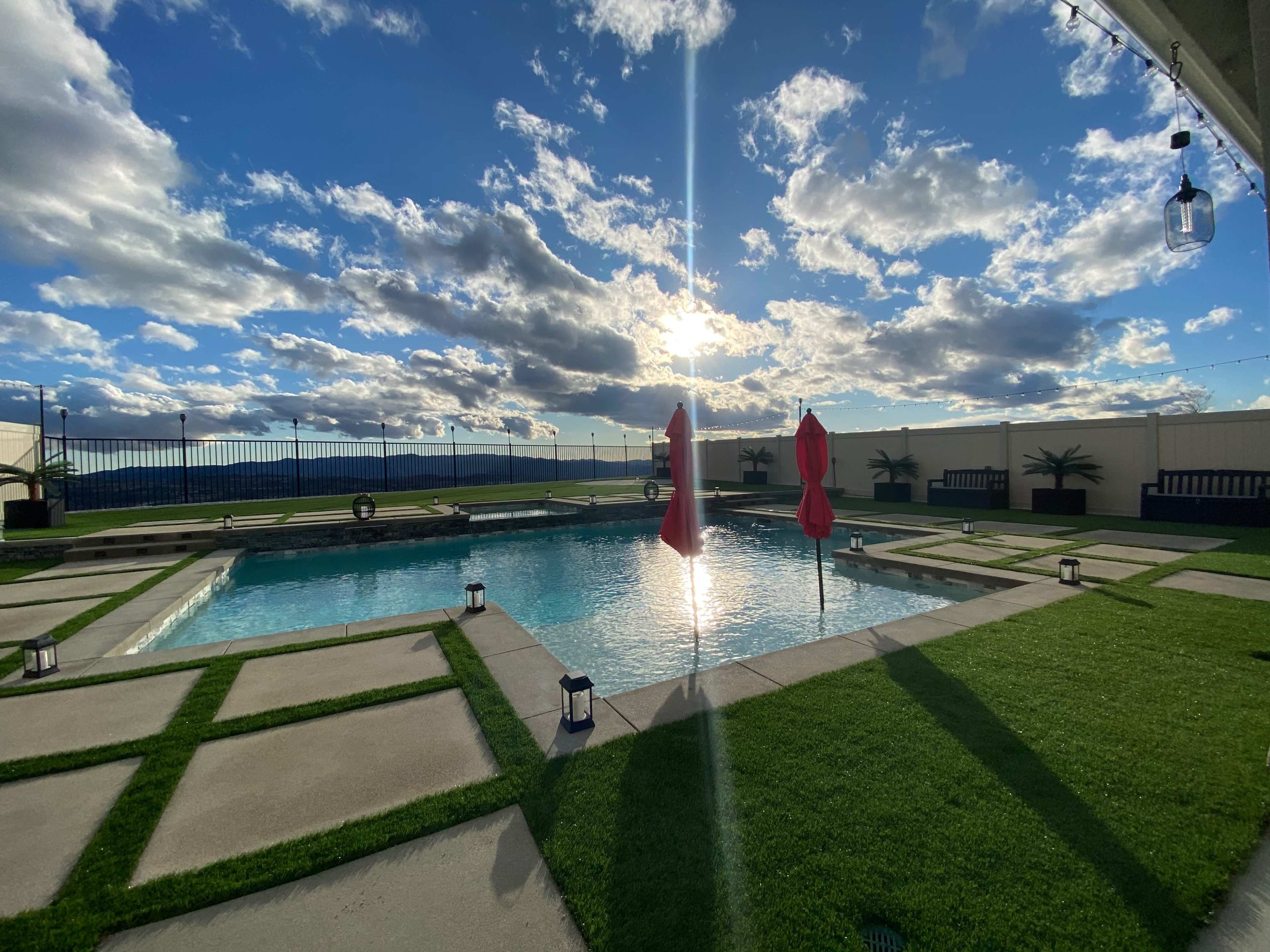 The image shows a landscaped pool area with two red umbrellas and a backdrop of cloudy skies at sunset.