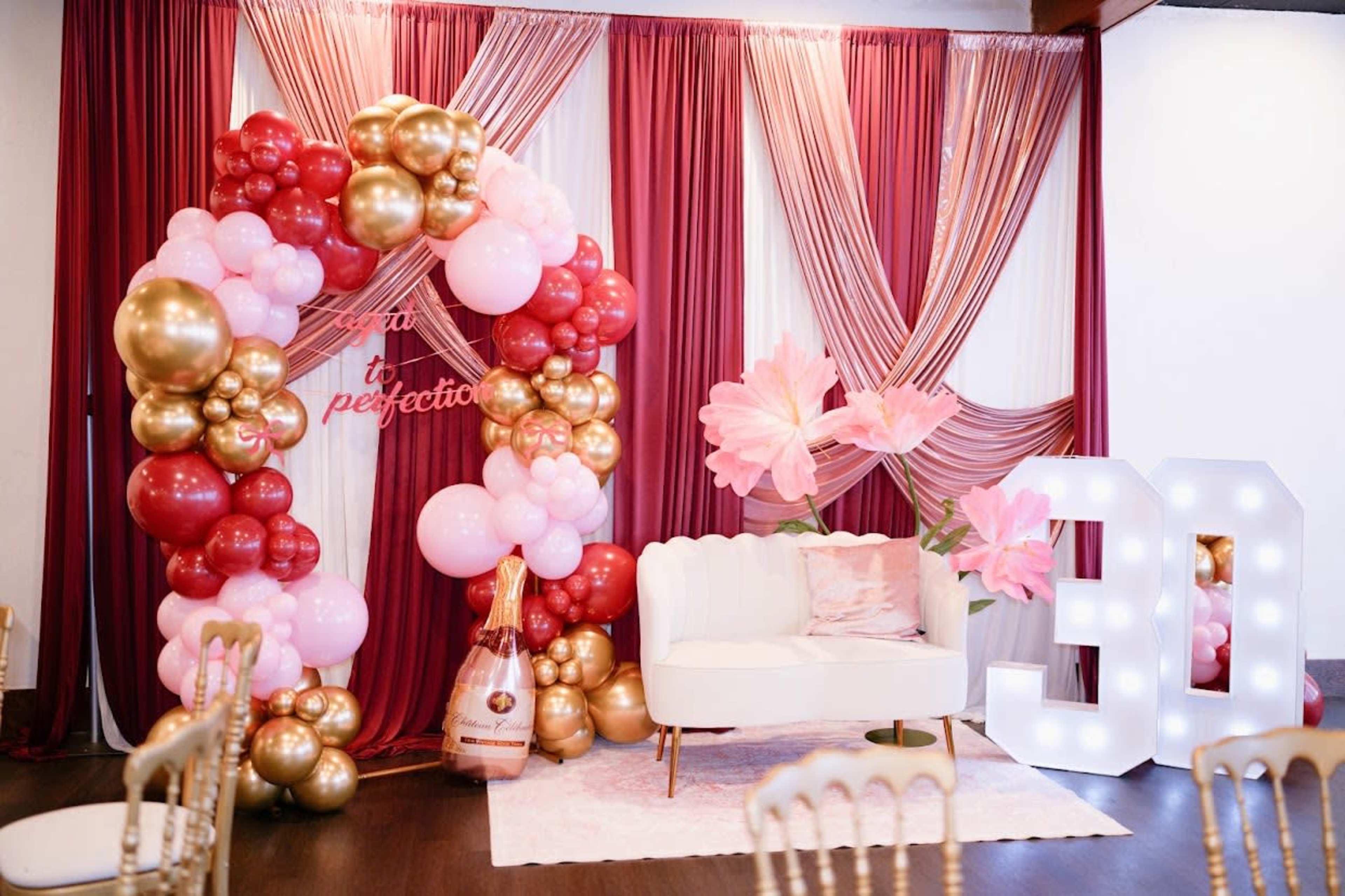 The image shows a decorated event space featuring a balloon arch in shades of pink, gold, and rose, alongside a white sofa and illuminated 30 sign.