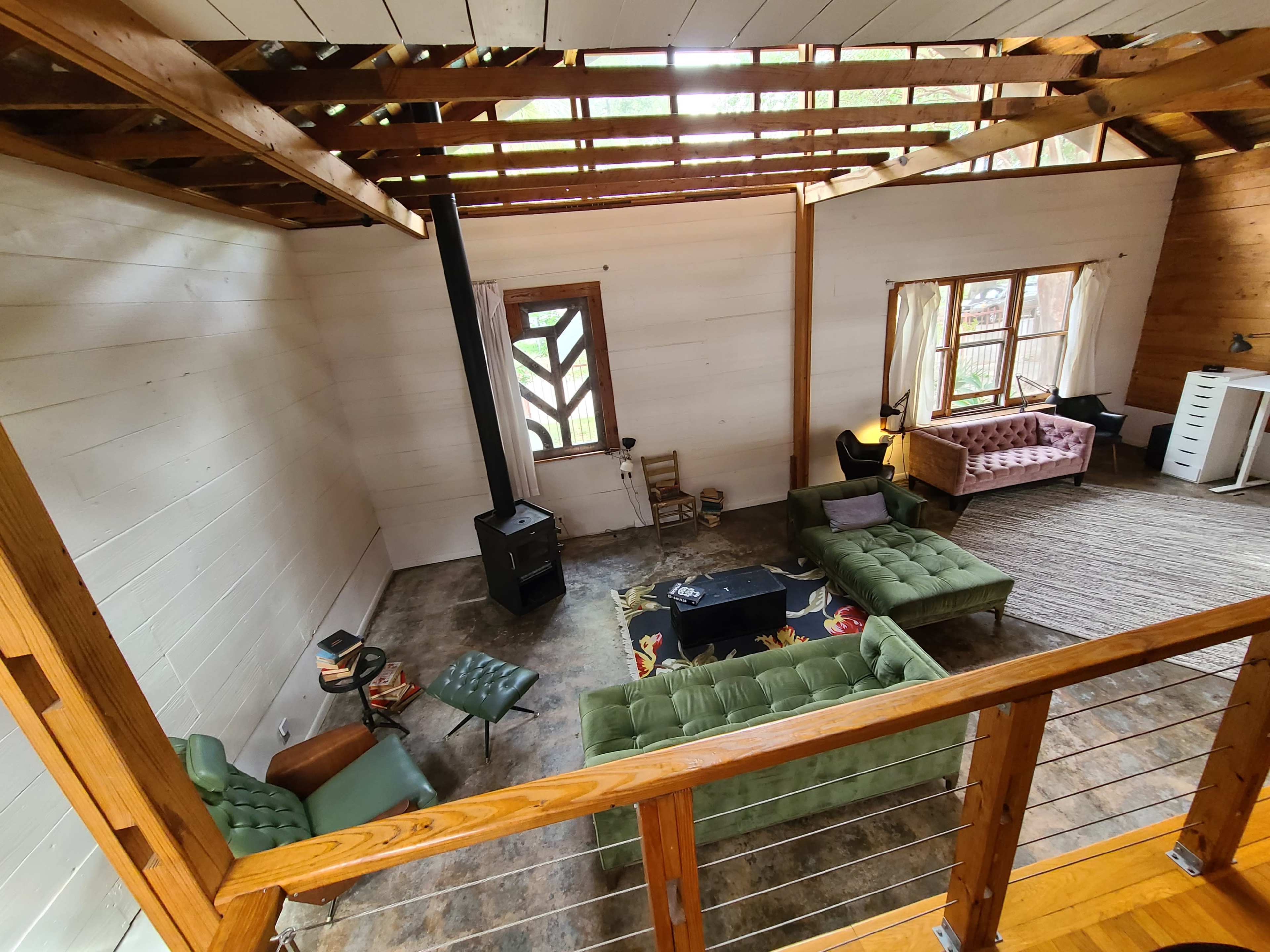 The image shows a spacious living area with green sofas, a wood stove, and large windows, featuring a mix of wood and concrete flooring.