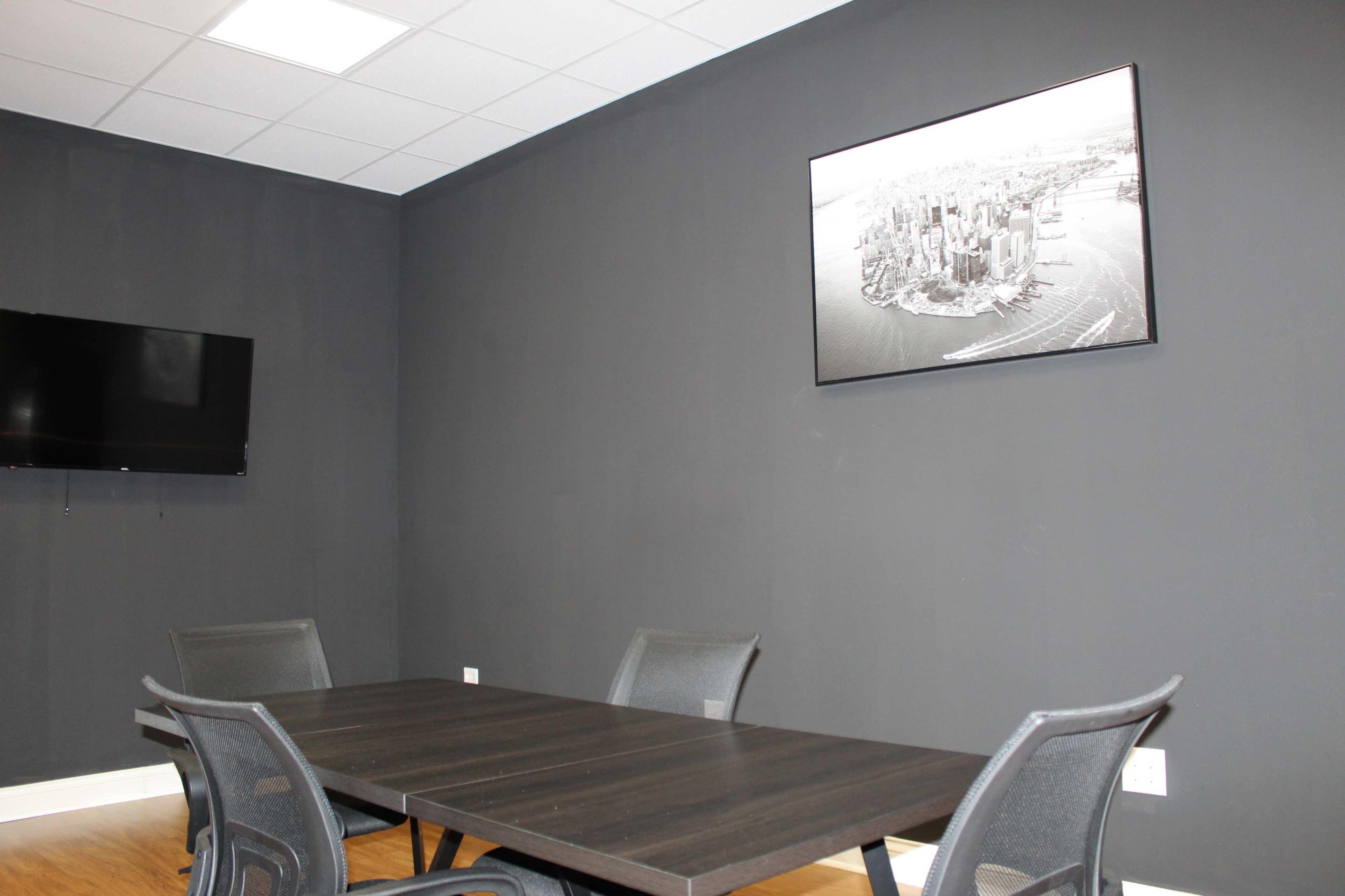 A conference room features a dark wall, a rectangular table surrounded by four chairs, a television mounted on one wall, and a framed black-and-white photograph of a cityscape above the table.
