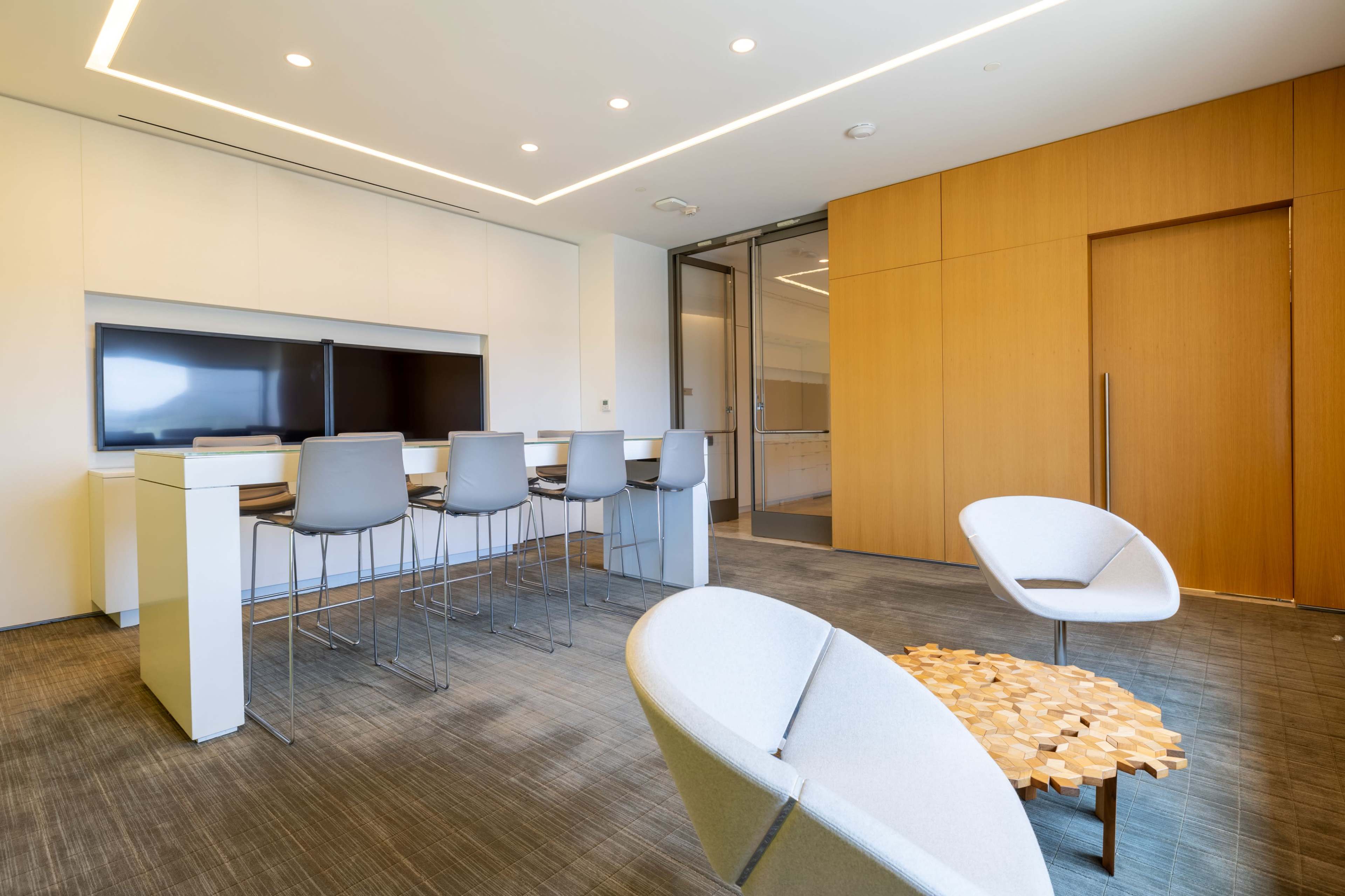 The image shows a modern office space featuring a bar-style seating area with several stools, a table made of wooden blocks, and two lounge chairs.