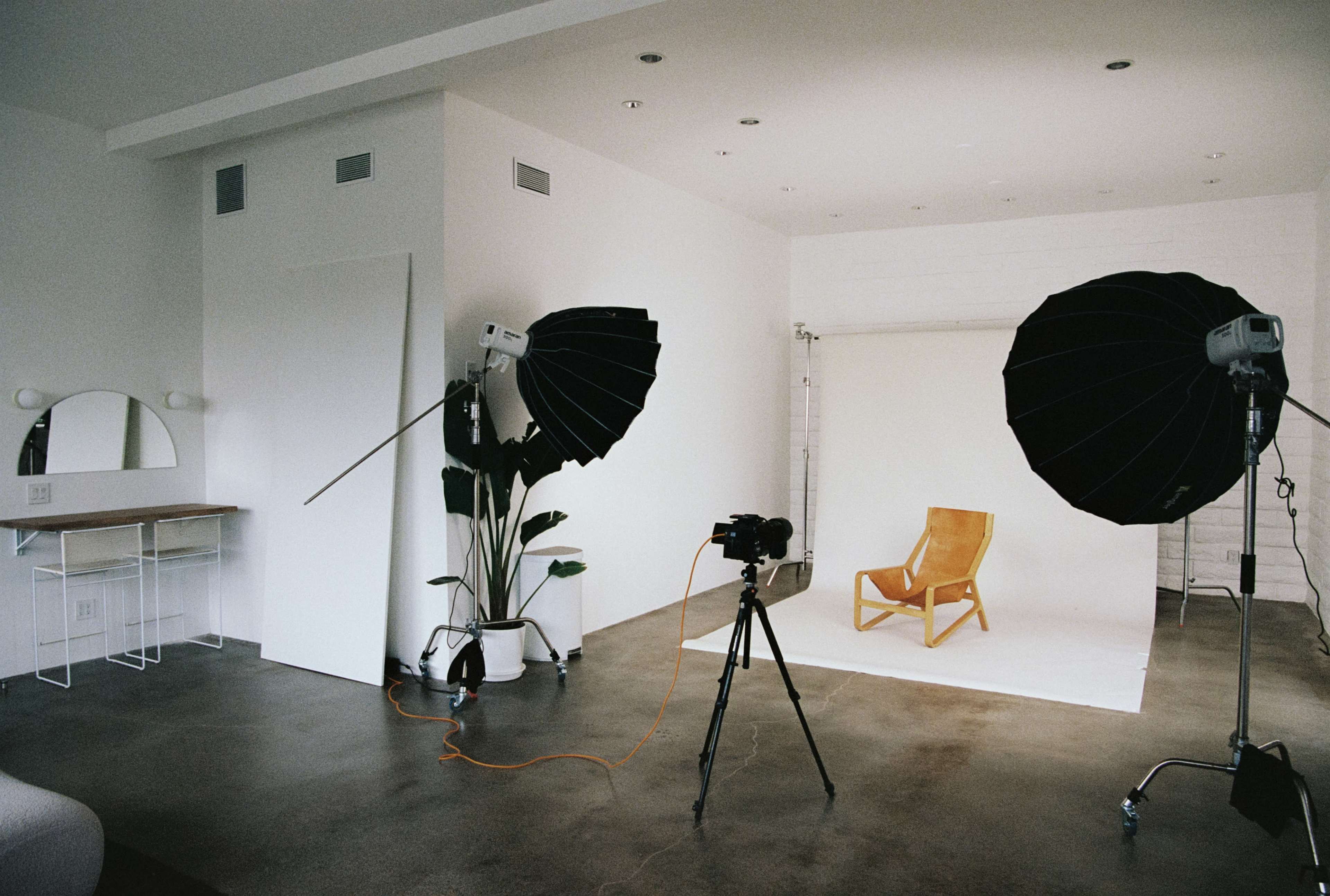 The image shows a photography studio with a wooden chair on a white backdrop, flanked by two large softbox lights and a camera on a tripod.