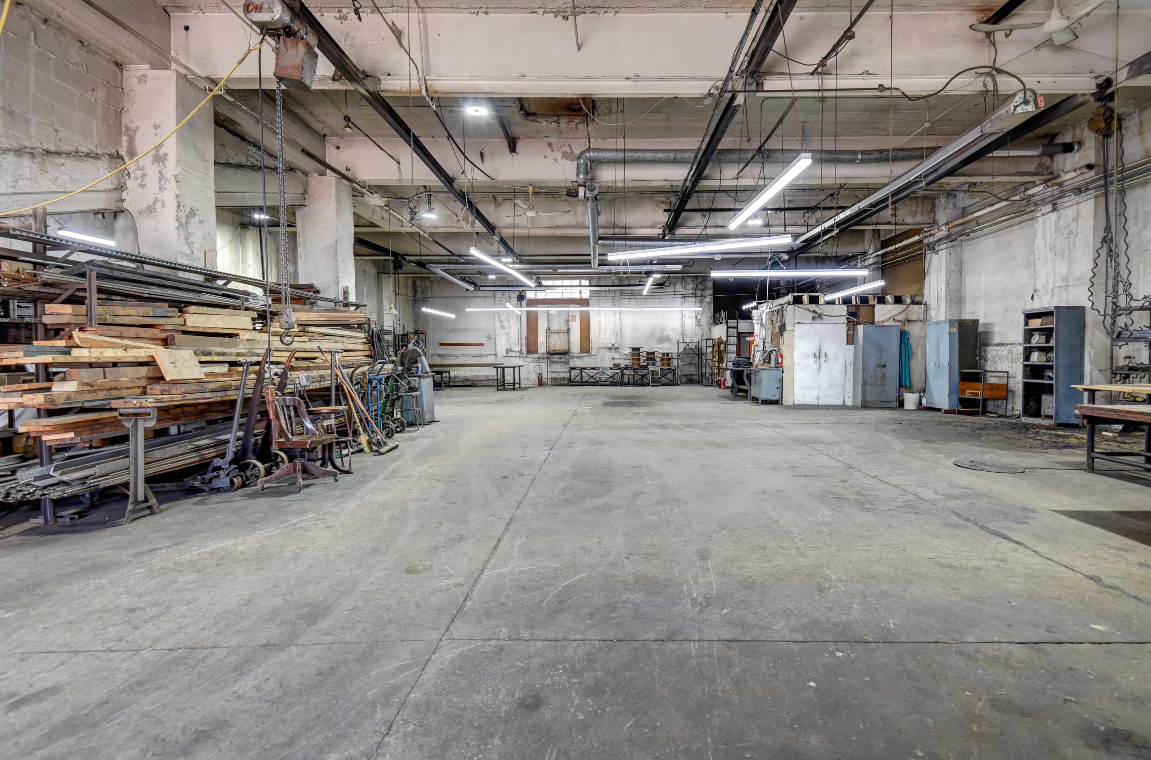 An empty industrial workshop features stacks of lumber, metalworking equipment, and overhead lighting.