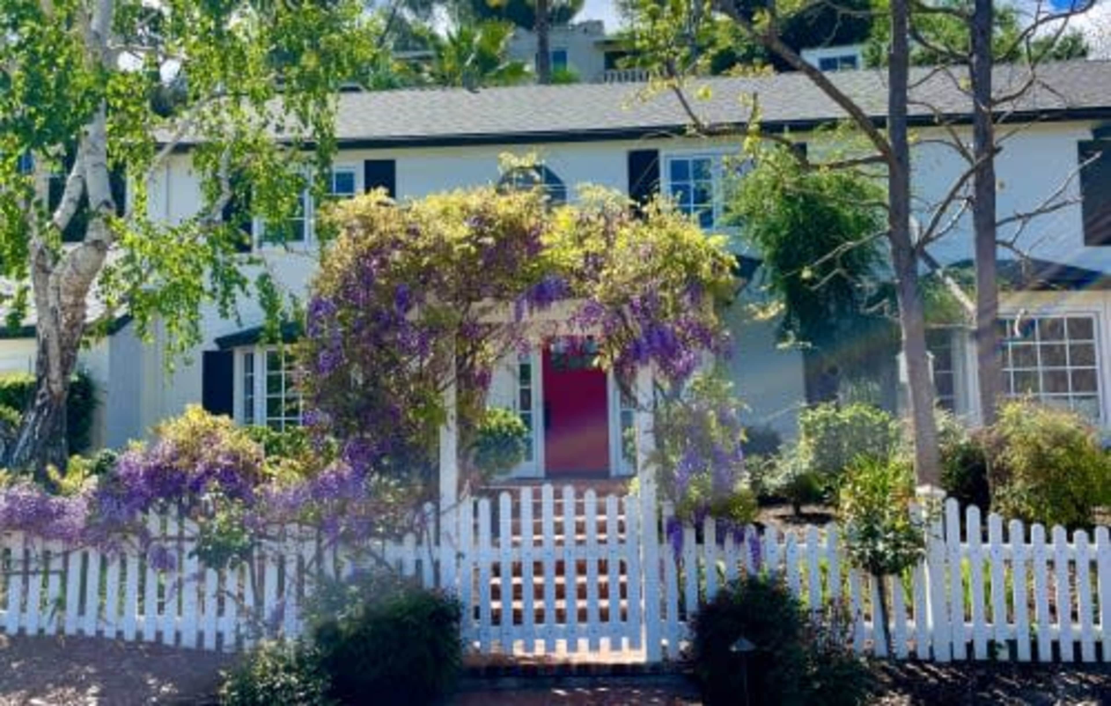 A two-story house with white walls, black shutters, and a vibrant entrance framed by purple flowers and a white picket fence.