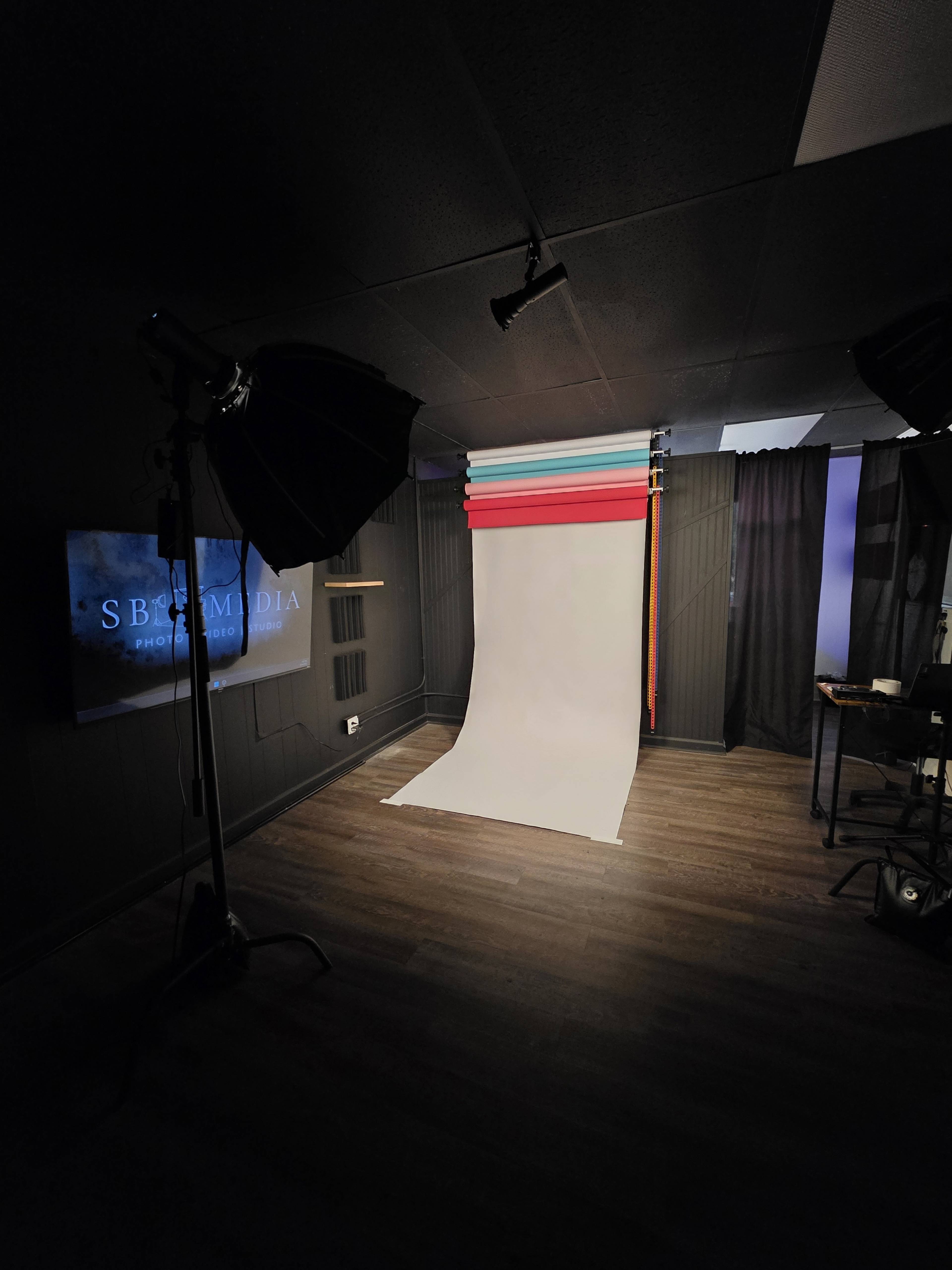 The image shows a photography studio with a white backdrop, colored paper rolls, softbox lights, and a viewing screen in a dimly lit room.
