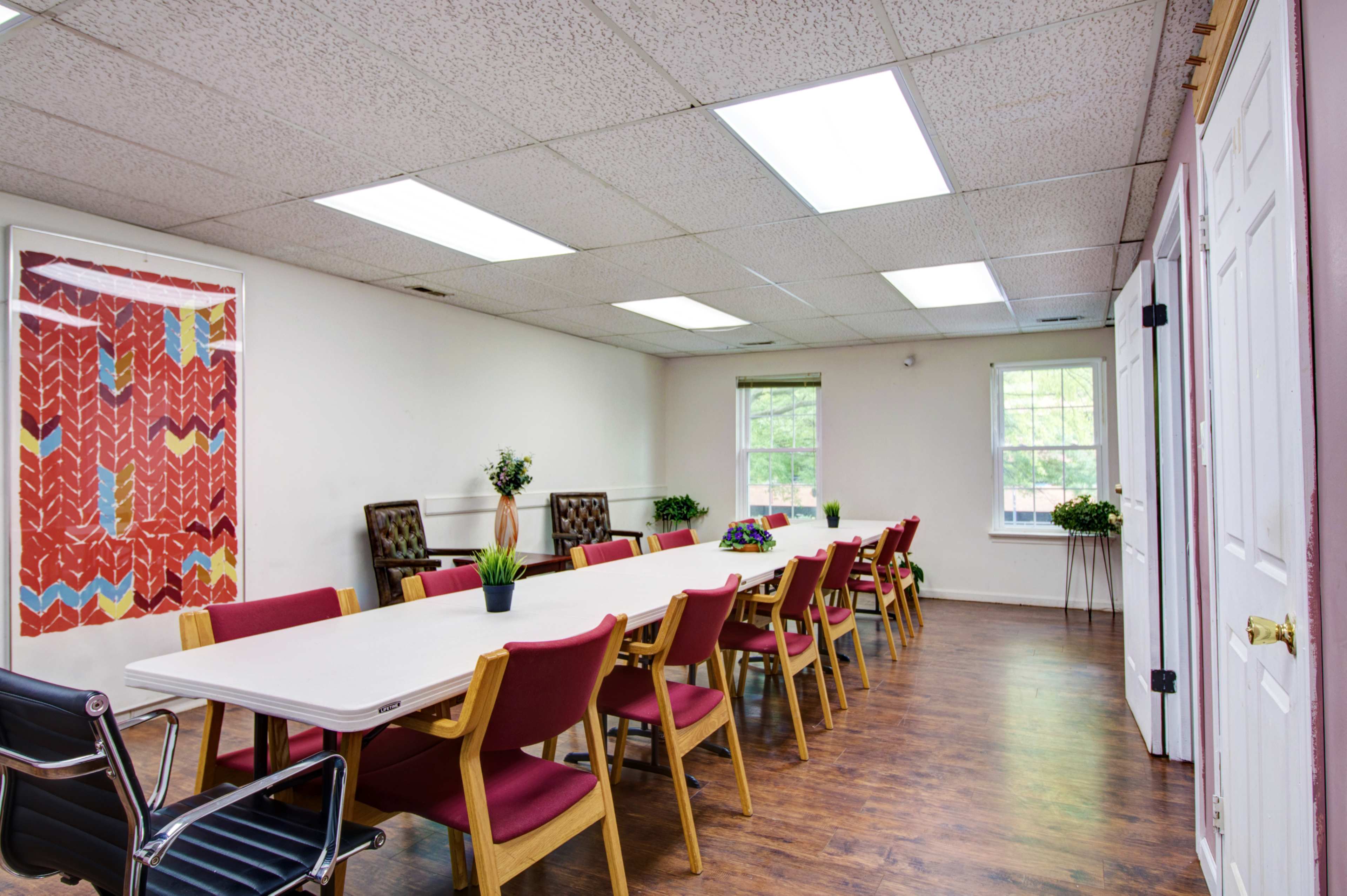 A spacious meeting room features a long white table surrounded by red chairs, with large overhead lights and windows providing ample illumination.