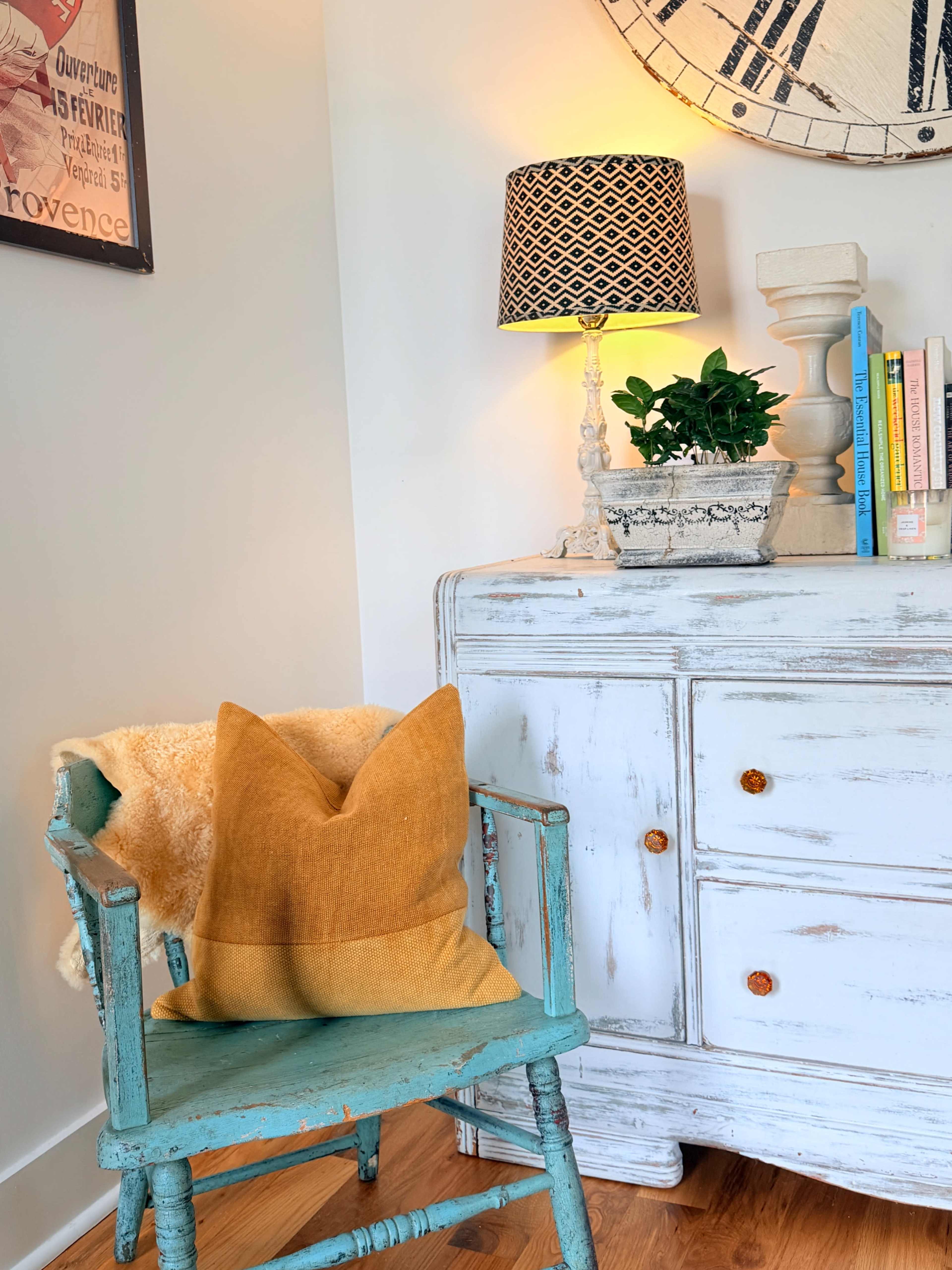 A vintage blue wooden chair with a mustard-colored pillow sits beside a white dresser adorned with a potted plant and a lamp.