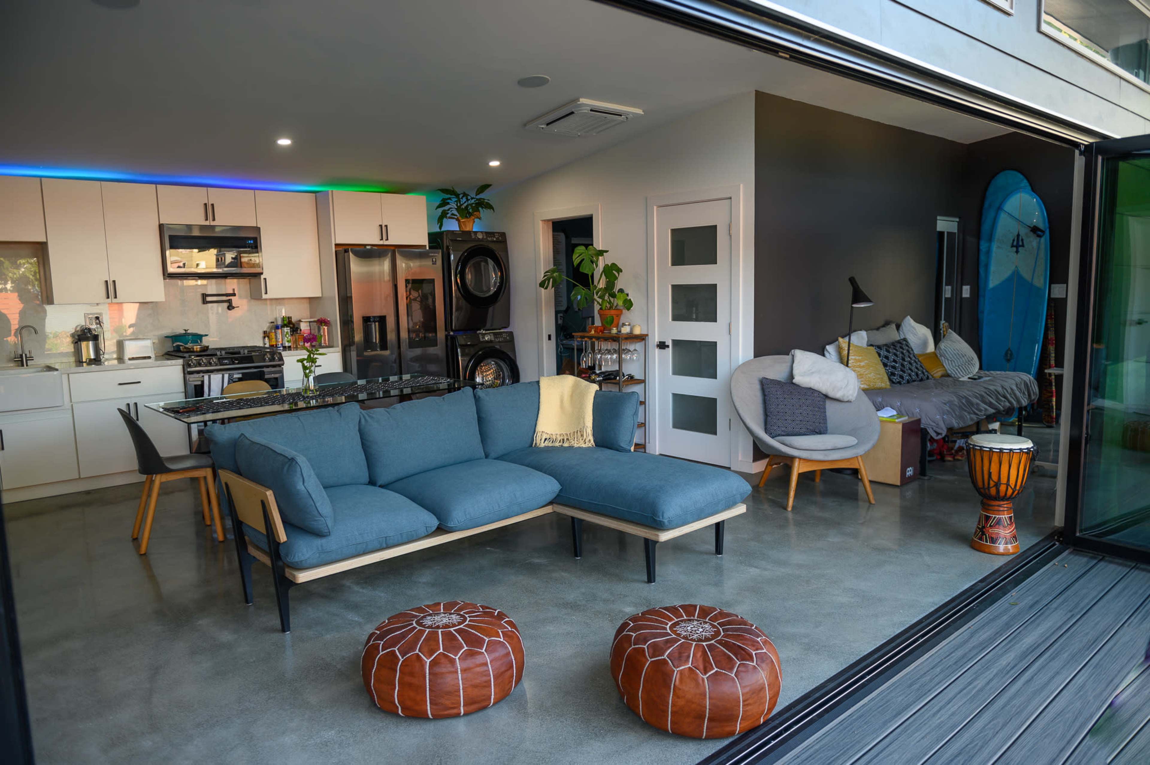 A modern living space features a blue sectional sofa, a round wooden drum, and two patterned ottomans, with an open kitchen and laundry area in the background.