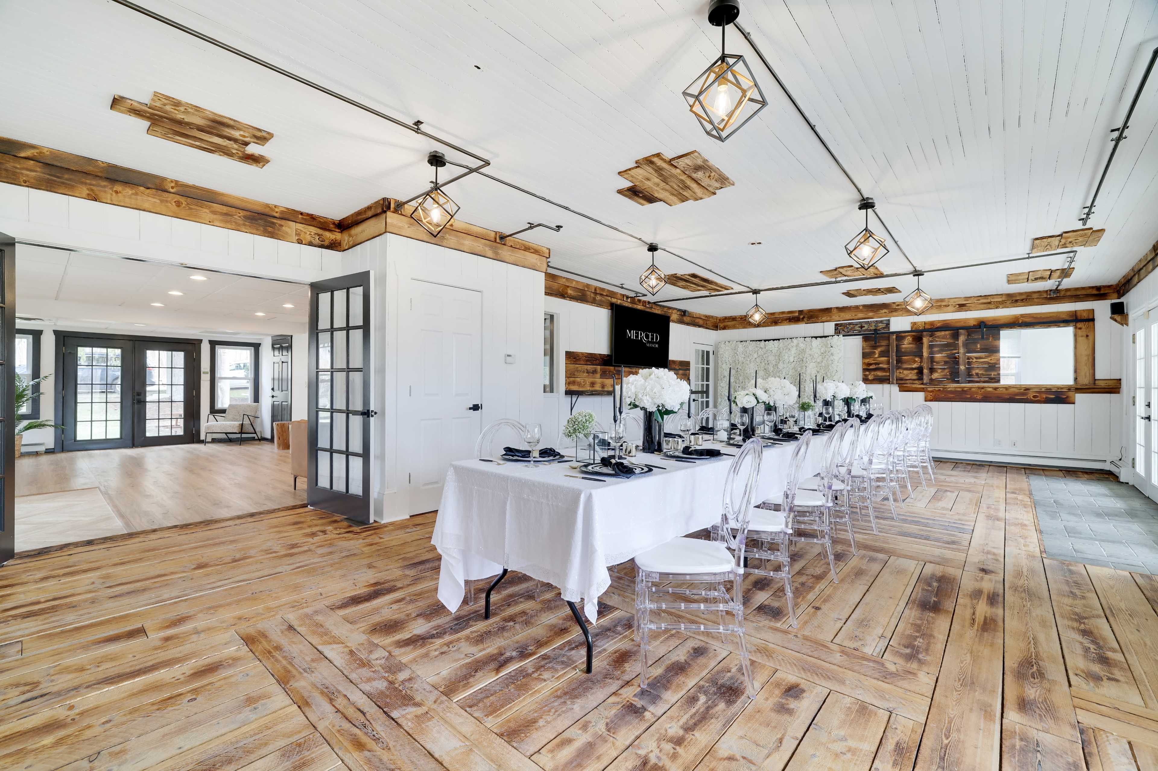 A spacious dining room with a long table set for a meal, surrounded by clear chairs and featuring wooden floors and modern light fixtures.