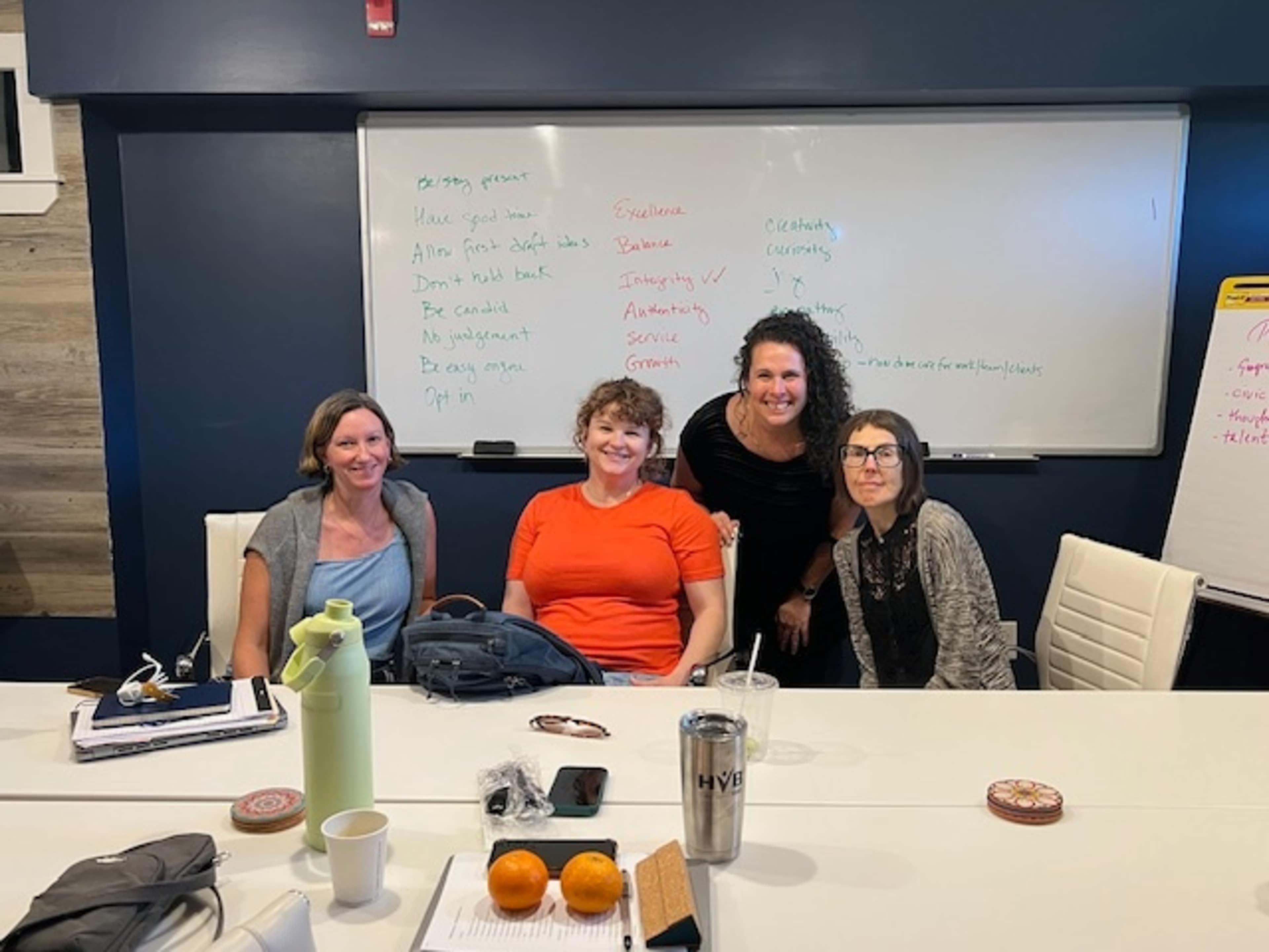 Four women are seated at a conference table in a meeting room, with a whiteboard in the background displaying notes and brainstorming ideas.