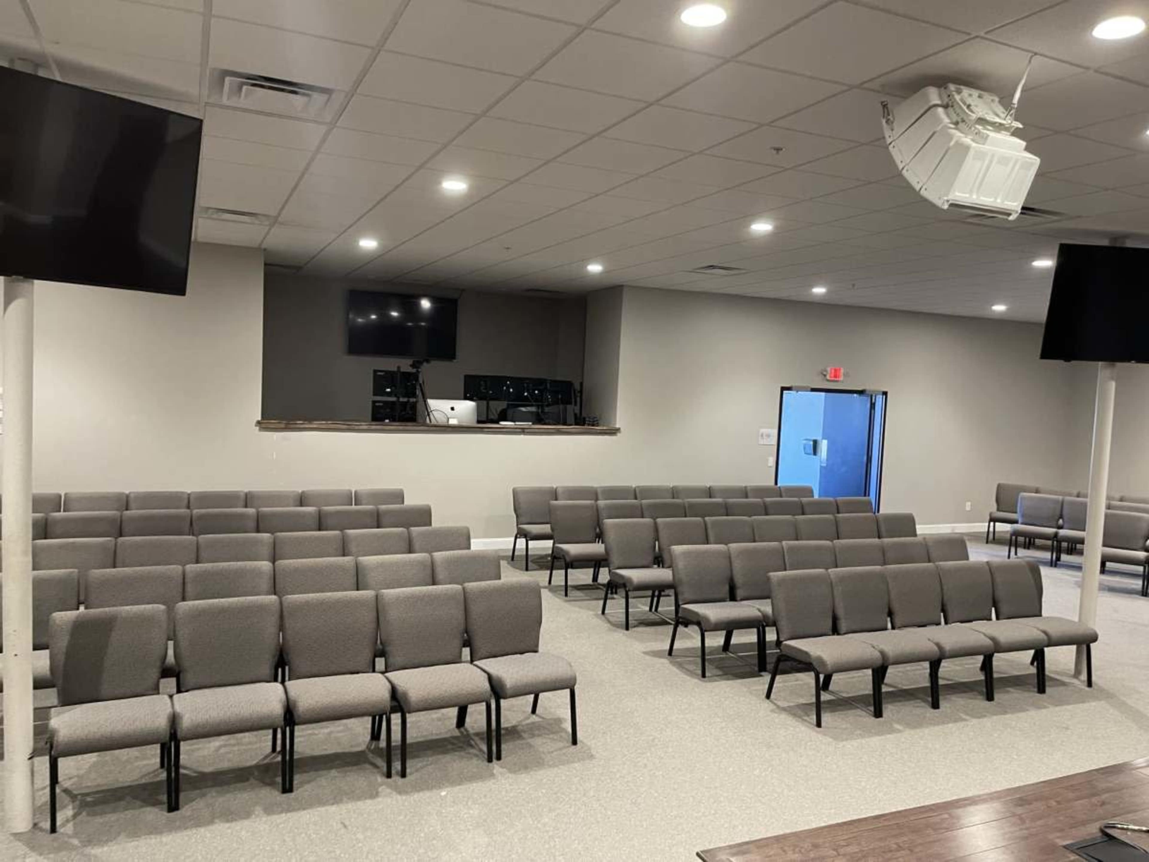 The image shows a large, empty room with rows of gray chairs and a sound system setup in the back, along with two screens mounted on the walls.
