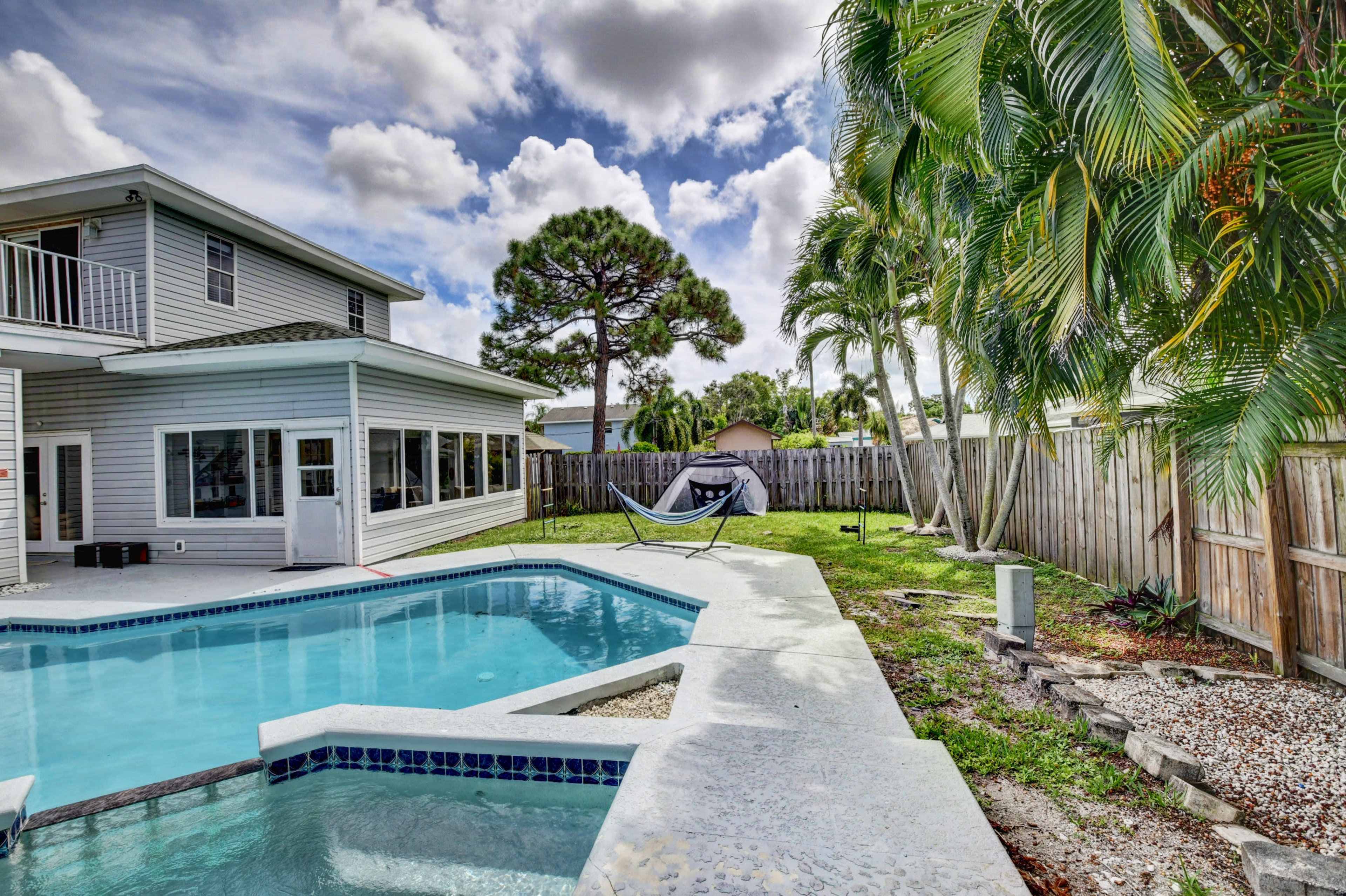 A backyard pool area with a hammock in a green lawn, surrounded by palm trees and a wooden fence.