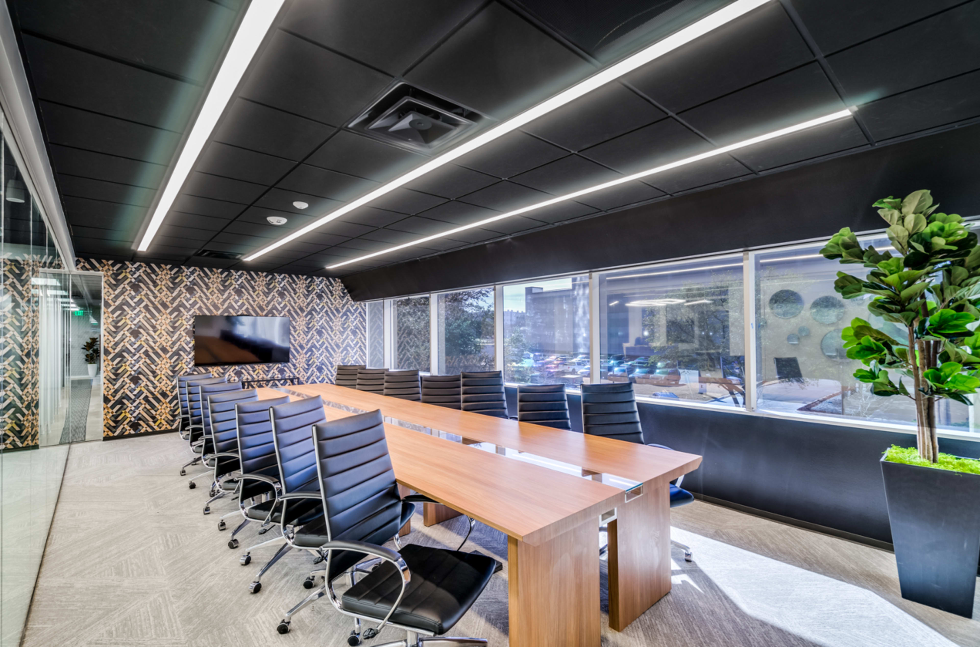 A modern conference room features a large wooden table surrounded by black ergonomic chairs, with large windows providing natural light and views of the outside.