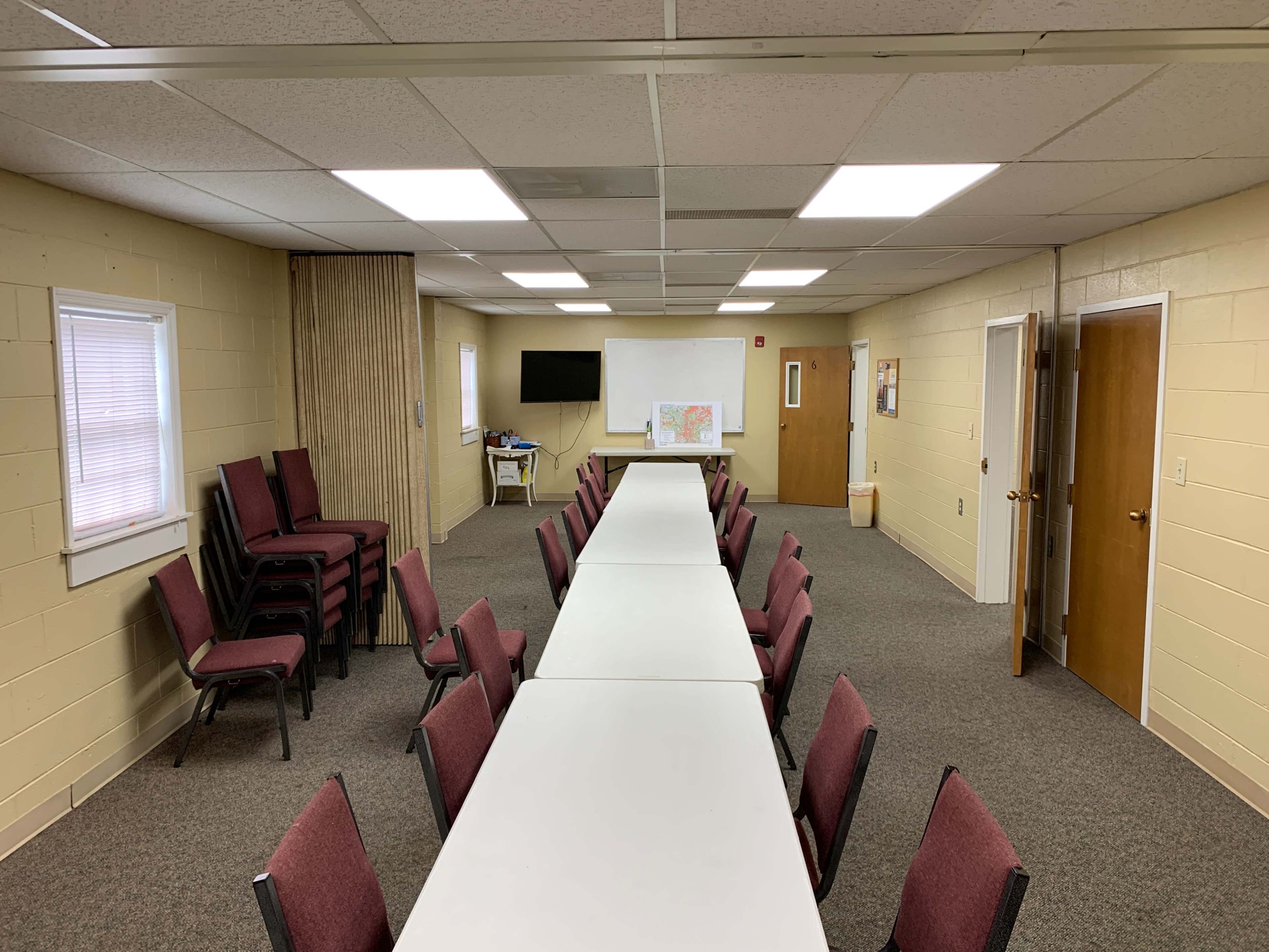 A long, rectangular table with chairs on either side is set up in a spacious room featuring beige walls, multiple light fixtures, and two doors.