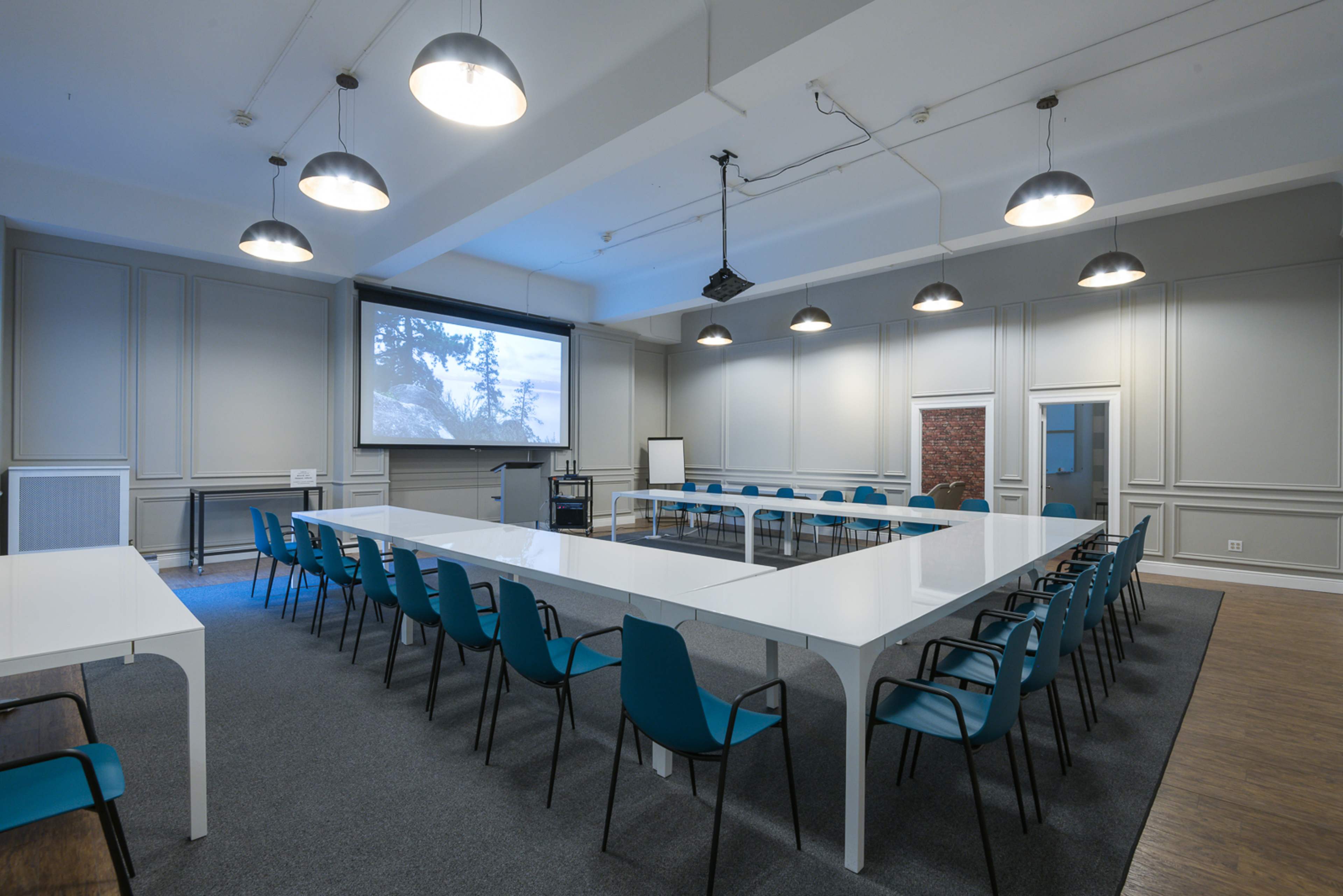 The image shows a modern meeting room featuring a long white table surrounded by turquoise chairs, with a projector screen at one end and various seating arrangements.