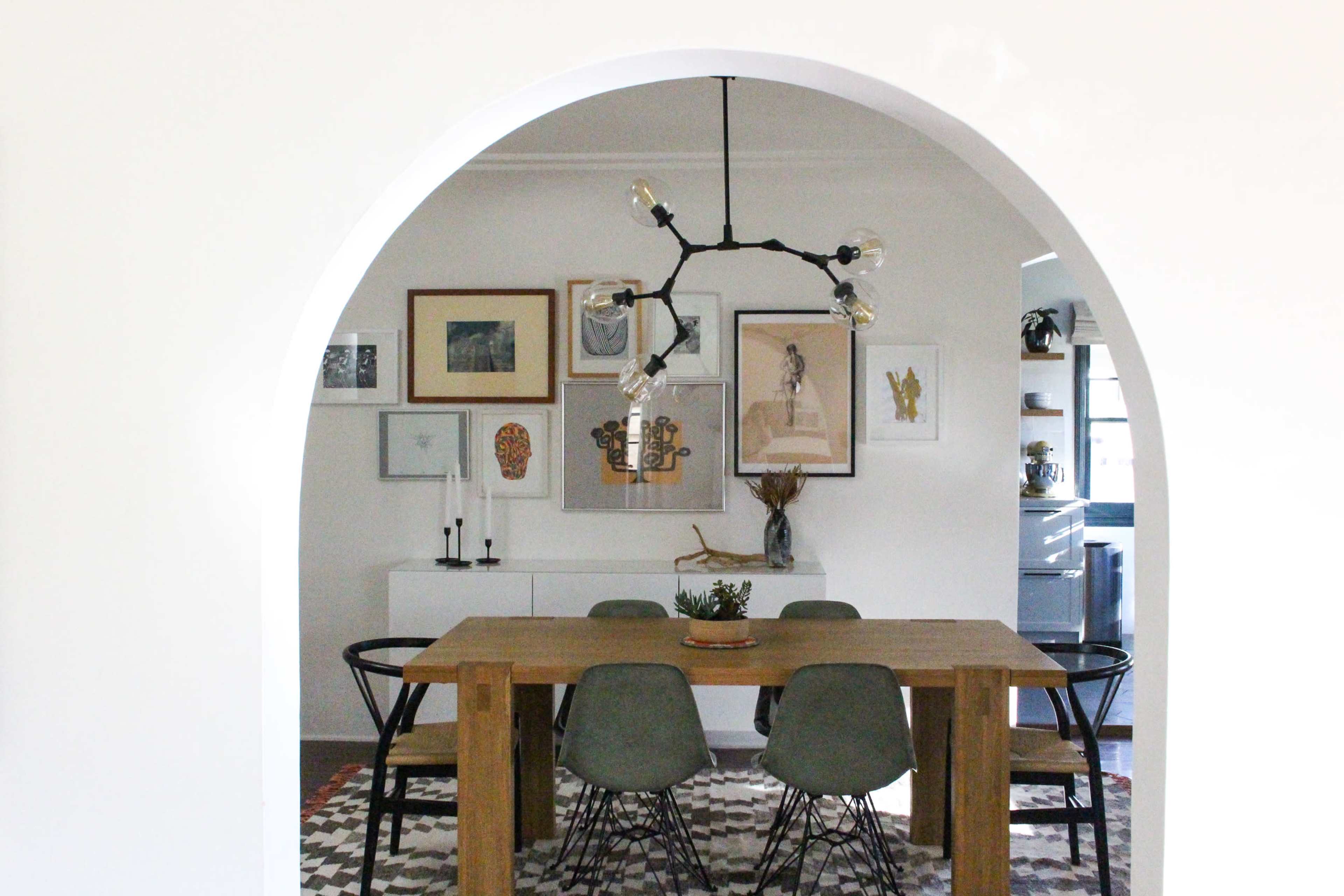 A wooden dining table surrounded by chairs sits under a modern chandelier in a room with framed artwork on the walls.