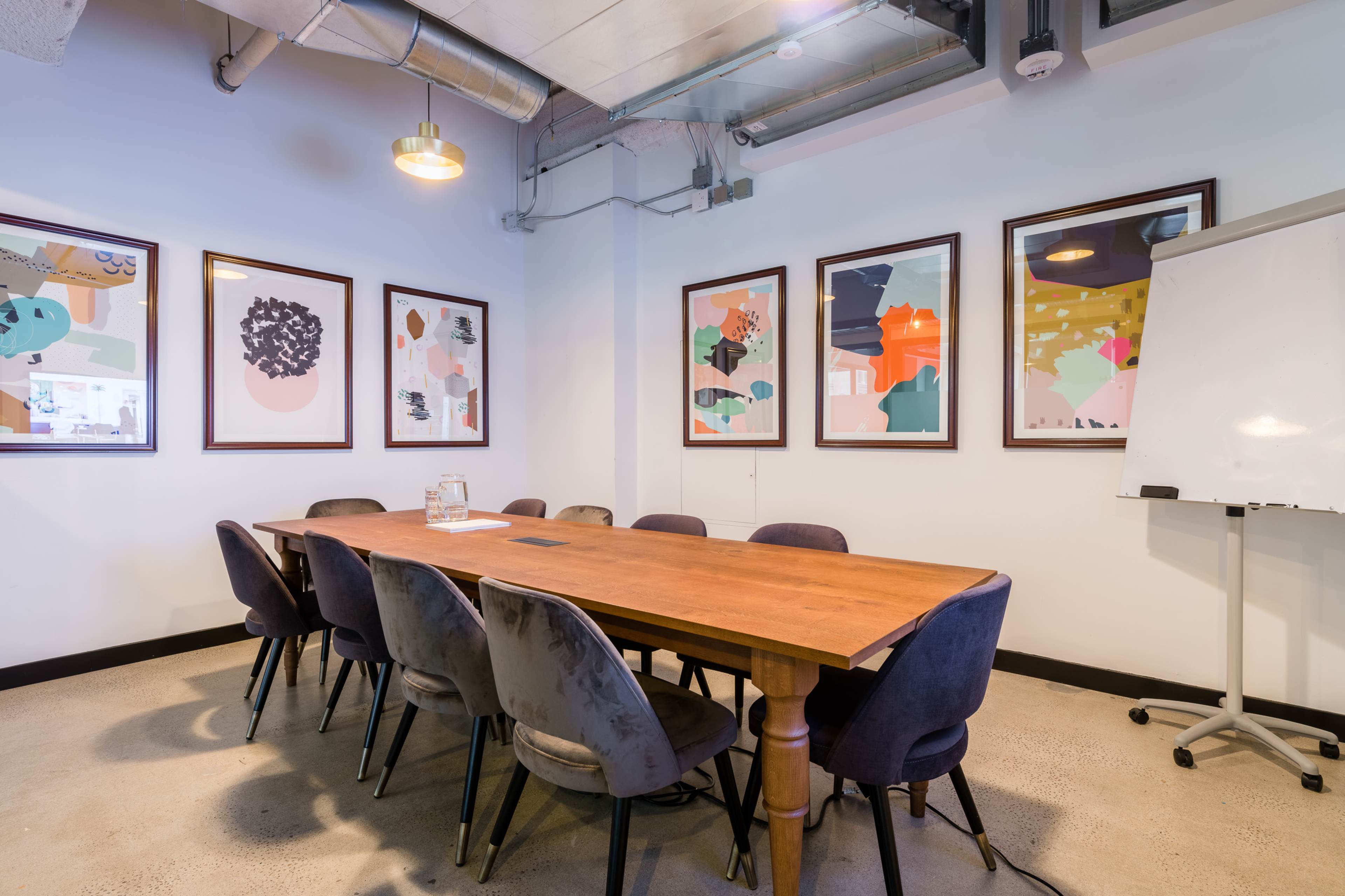 A long wooden conference table is surrounded by eight dark velvet chairs in a well-lit meeting room with framed abstract art on the walls.