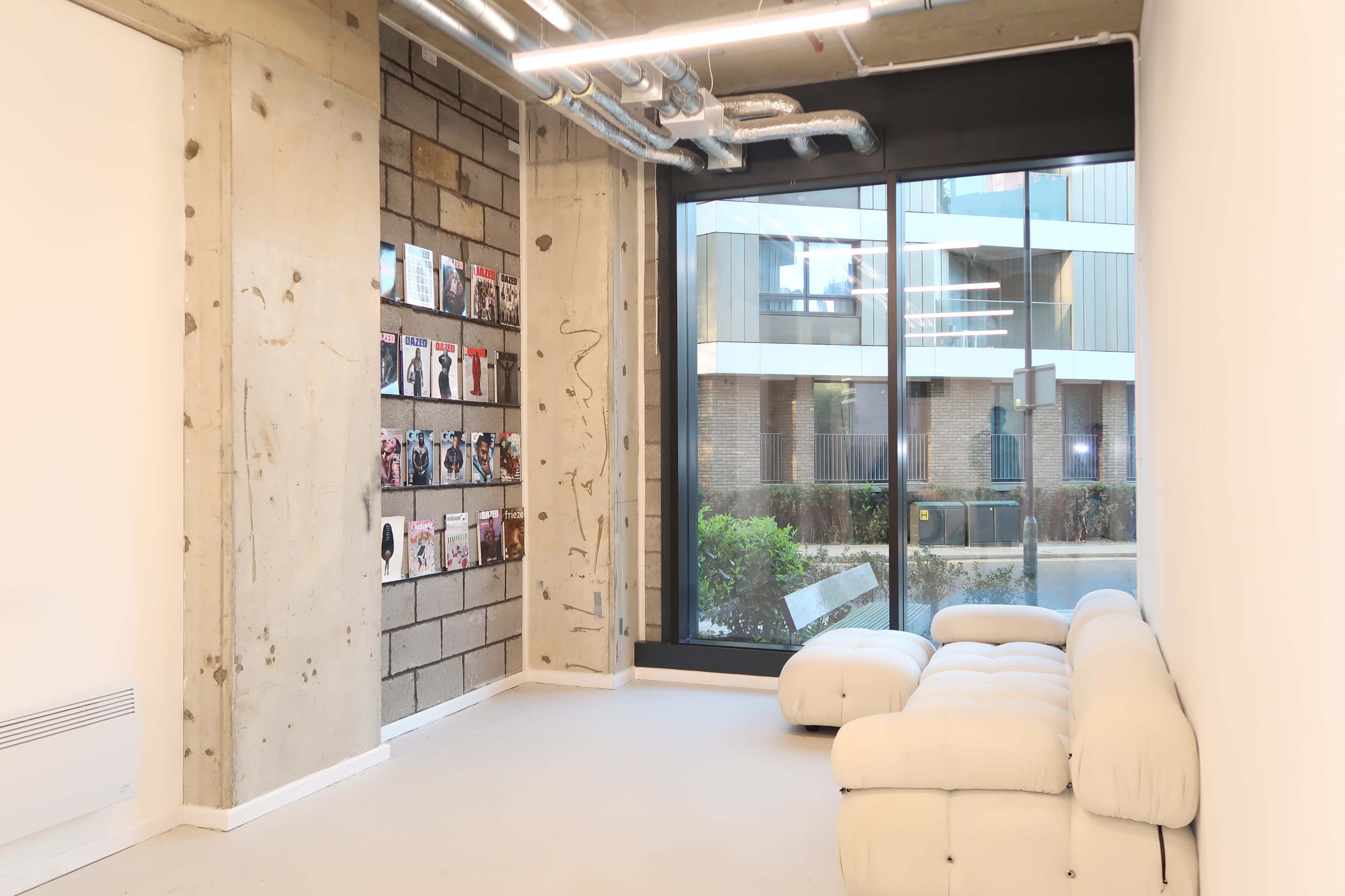The image shows a modern interior space featuring a white sectional sofa, exposed concrete walls, and a large window displaying a view of an outdoor area with green plants.