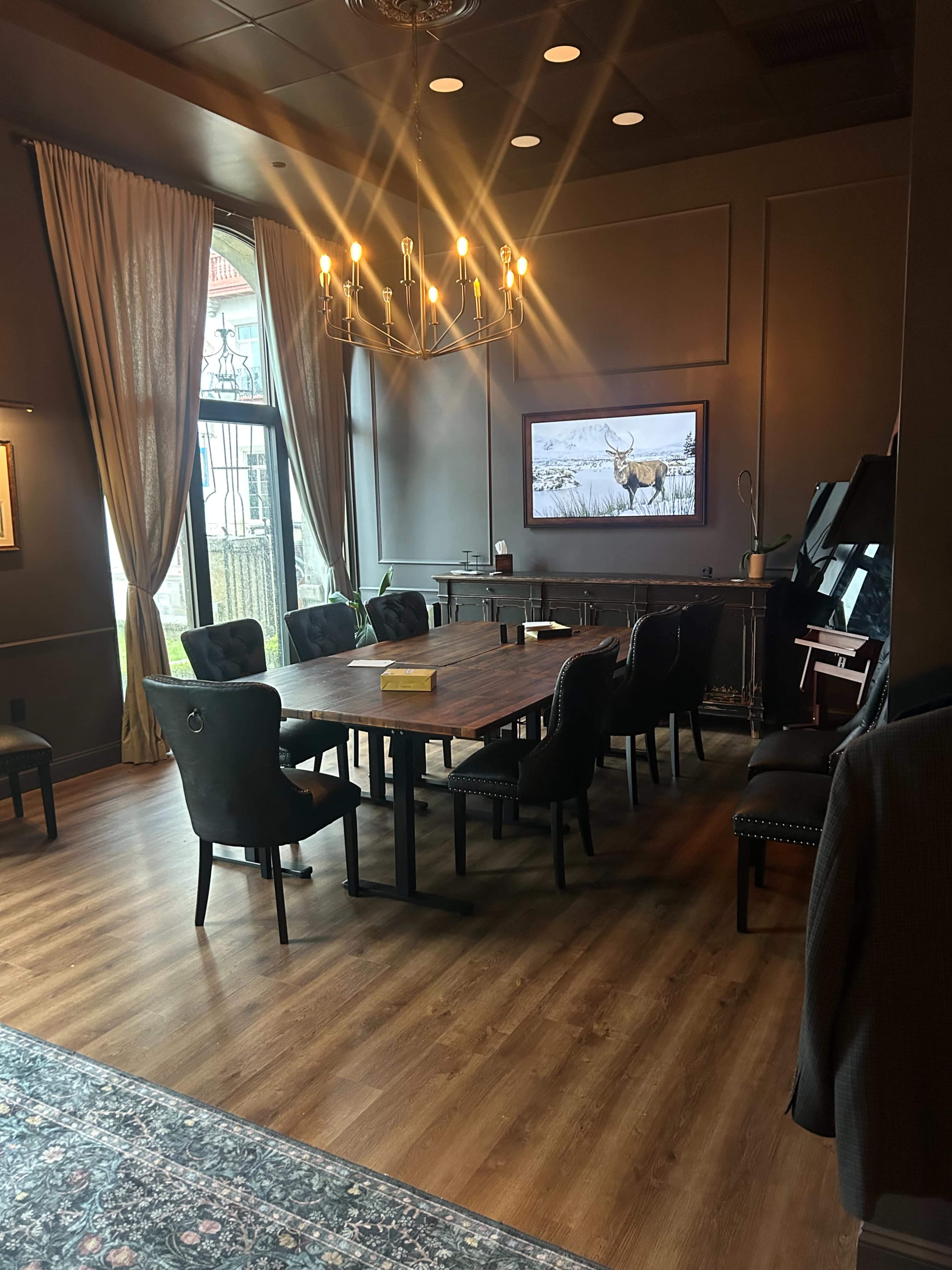 A large wooden table surrounded by black chairs is positioned in a well-lit meeting room featuring a window with a view and a large television on the wall.