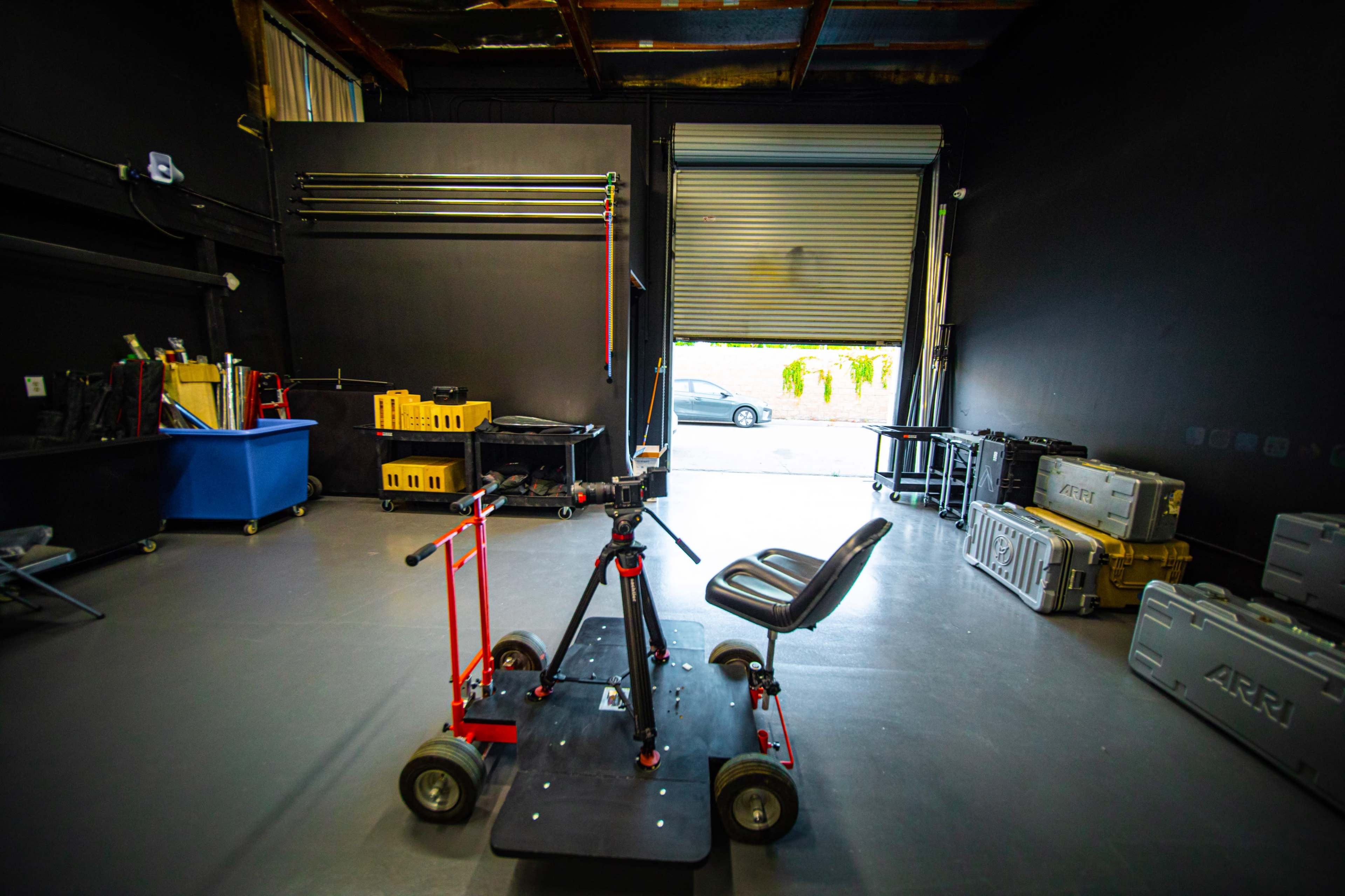 A camera dolly on wheels positioned in a dimly lit storage space with a large roll-up door partially open, revealing a car outside.