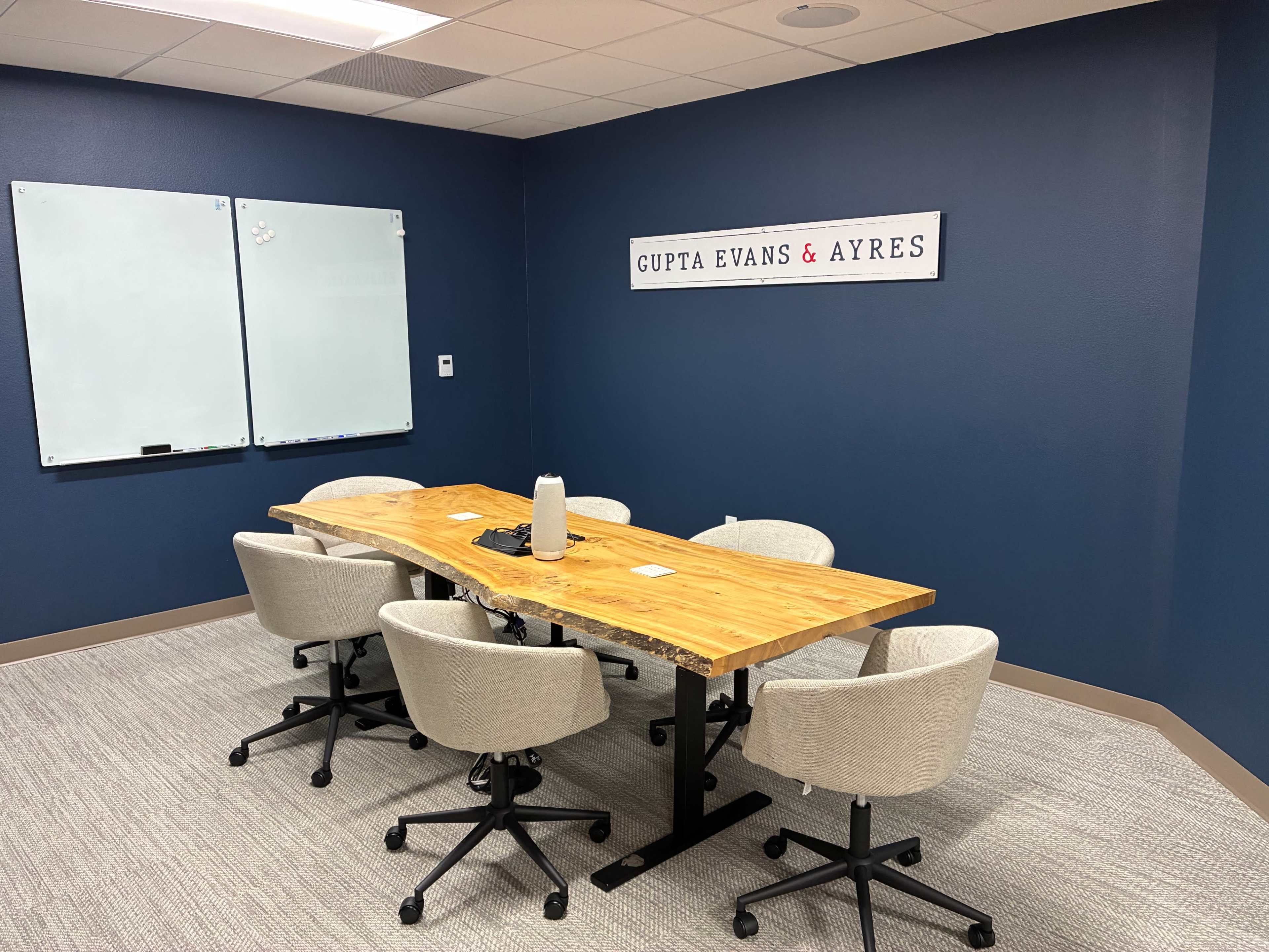 The image shows a conference room with a wooden table surrounded by five chairs, a whiteboard on one wall, and a sign reading "GUPTA EVANS & AYRES" on another wall.