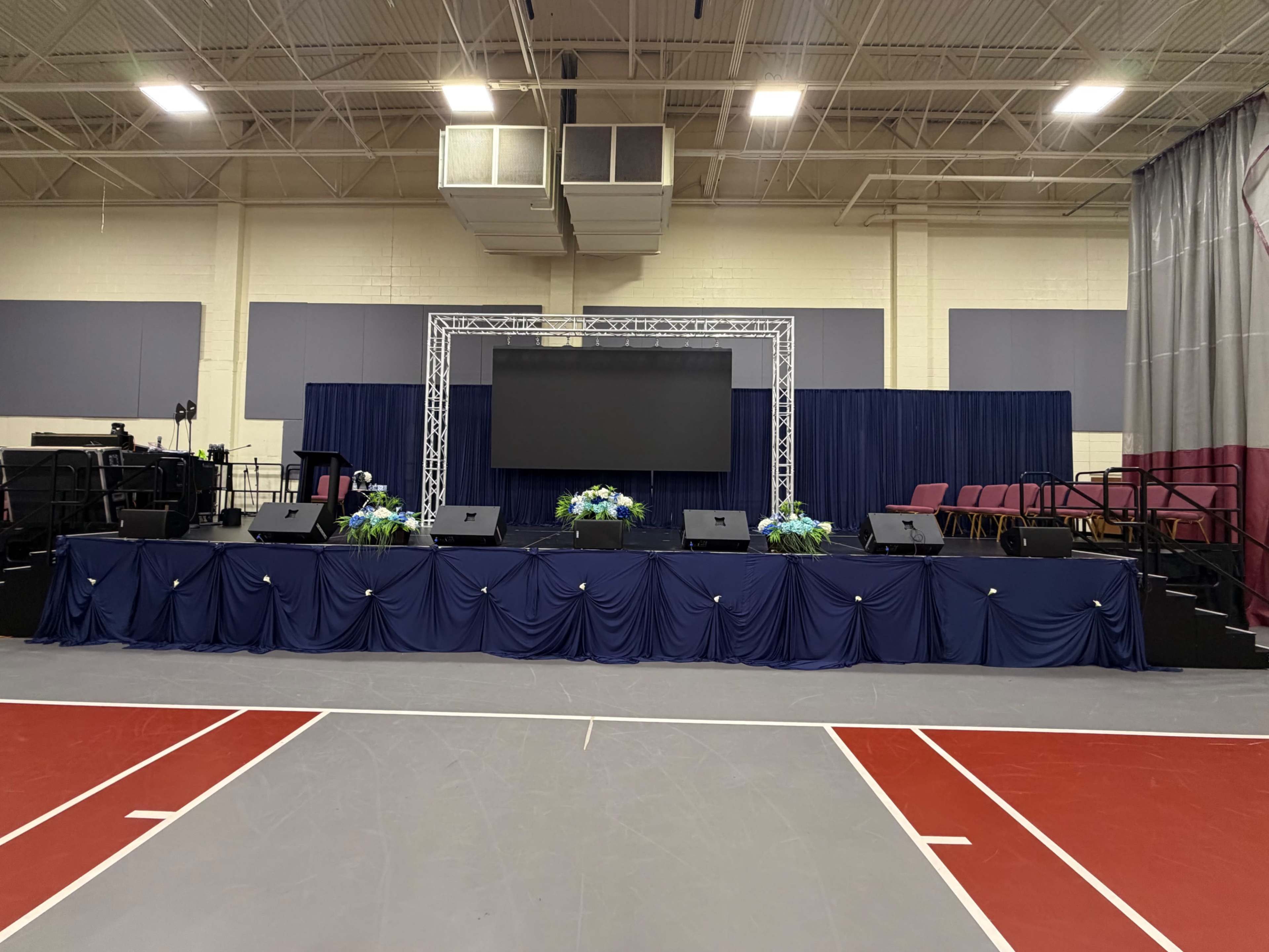 A stage setup with a large screen, podiums, and floral arrangements is prepared in a gymnasium.