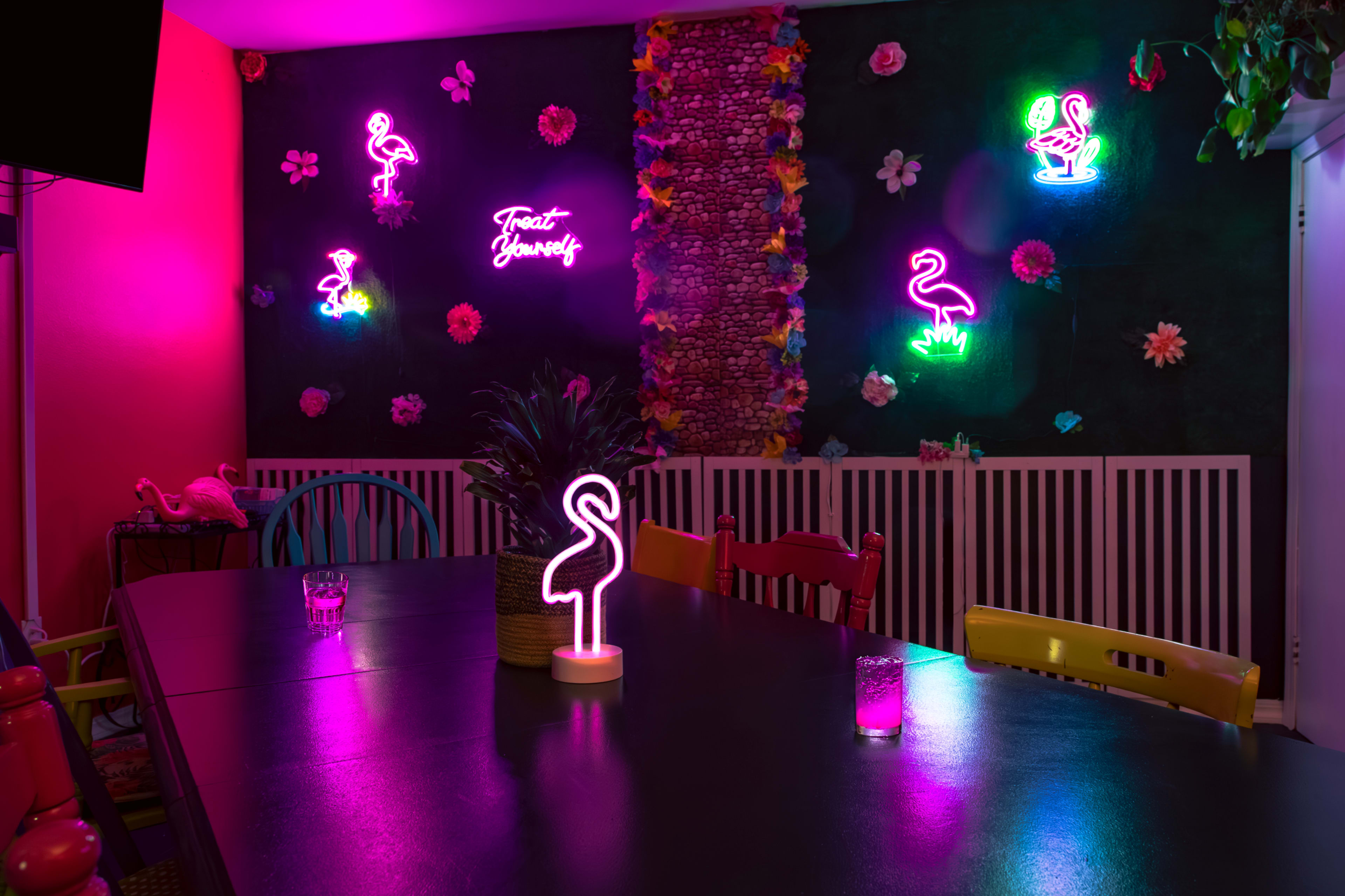 A brightly lit room features neon flamingo signs and floral decorations along the walls, with a large dark table and colorful chairs arranged around it.