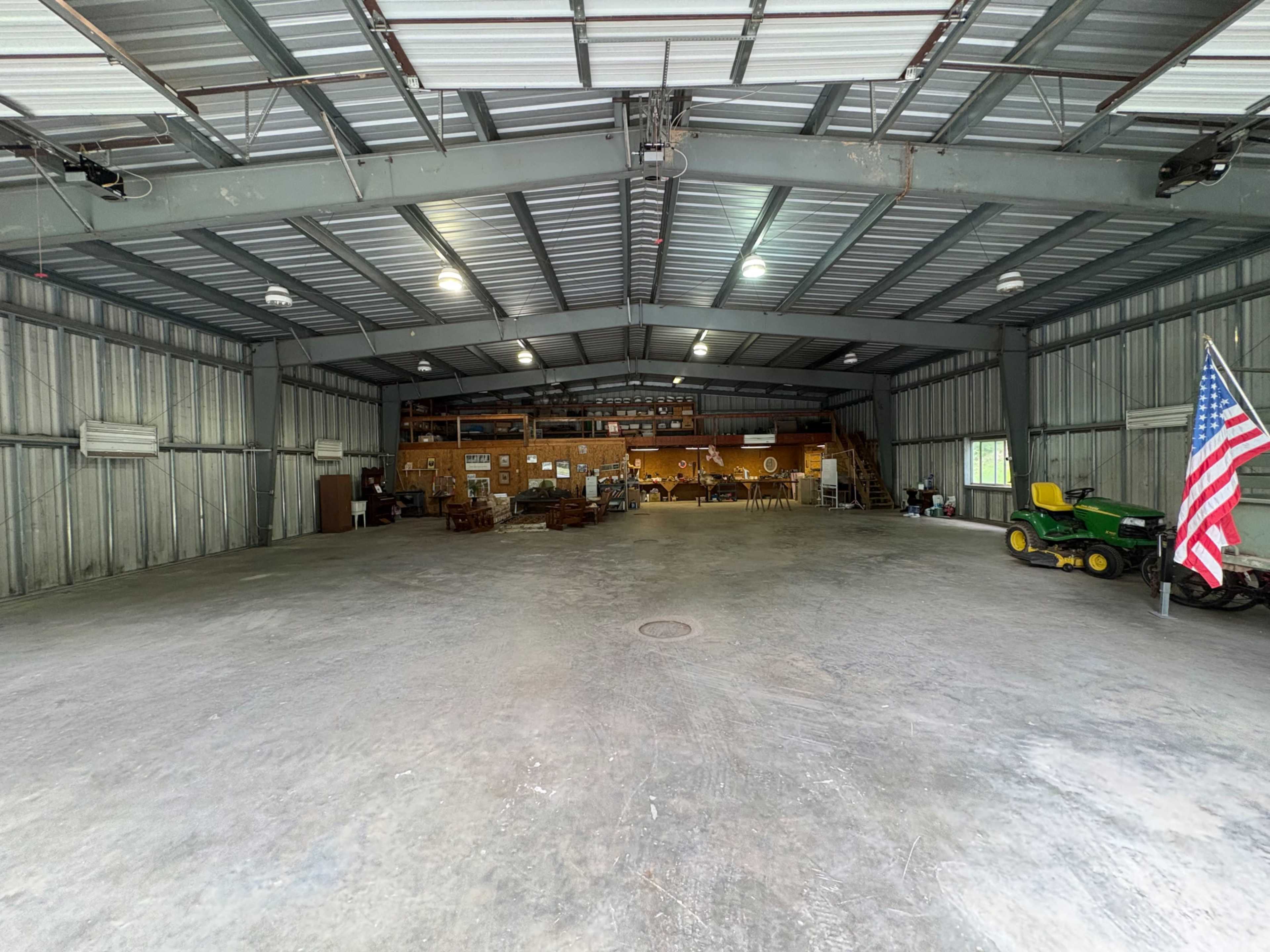 The image shows a large, open metal barn or warehouse space with concrete flooring, featuring various pieces of equipment and furniture along the sides, including a green tractor and an American flag.