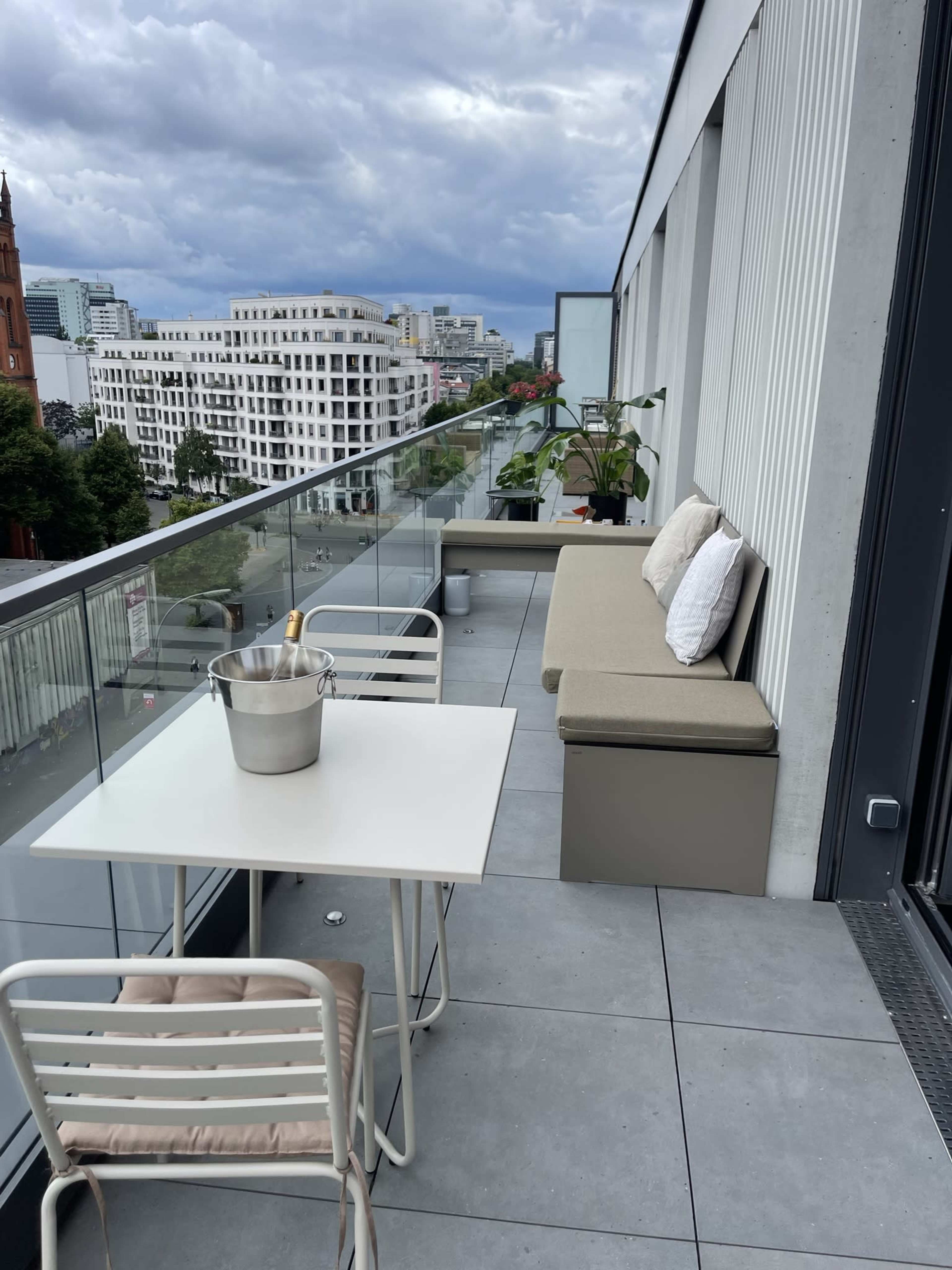 A balcony with a table and chairs, a bench, a planter, and a bucket, overlooking a cityscape under a cloudy sky.