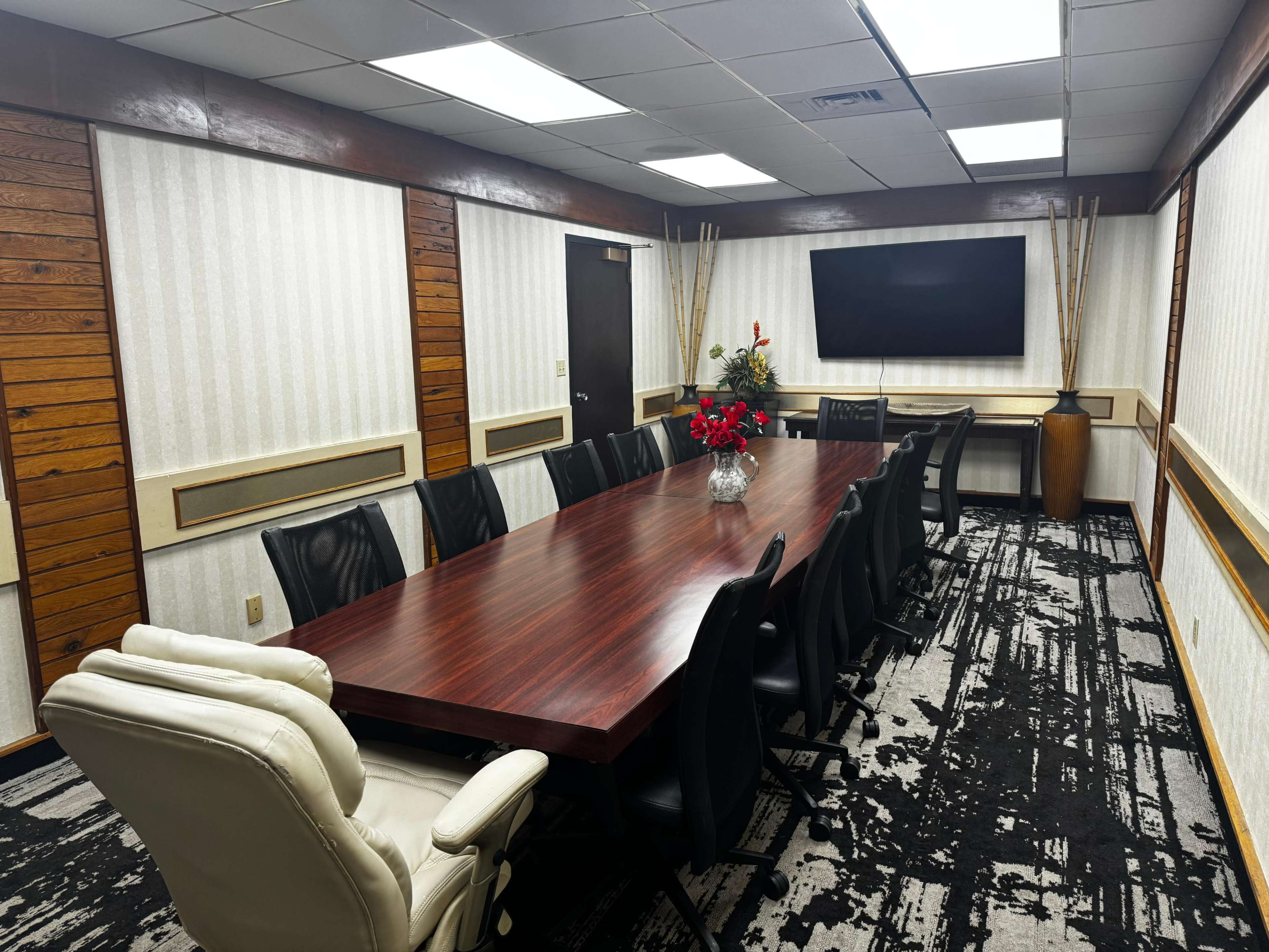 The image shows a conference room with a long wooden table surrounded by black chairs, a wall-mounted television, and decorative plants.