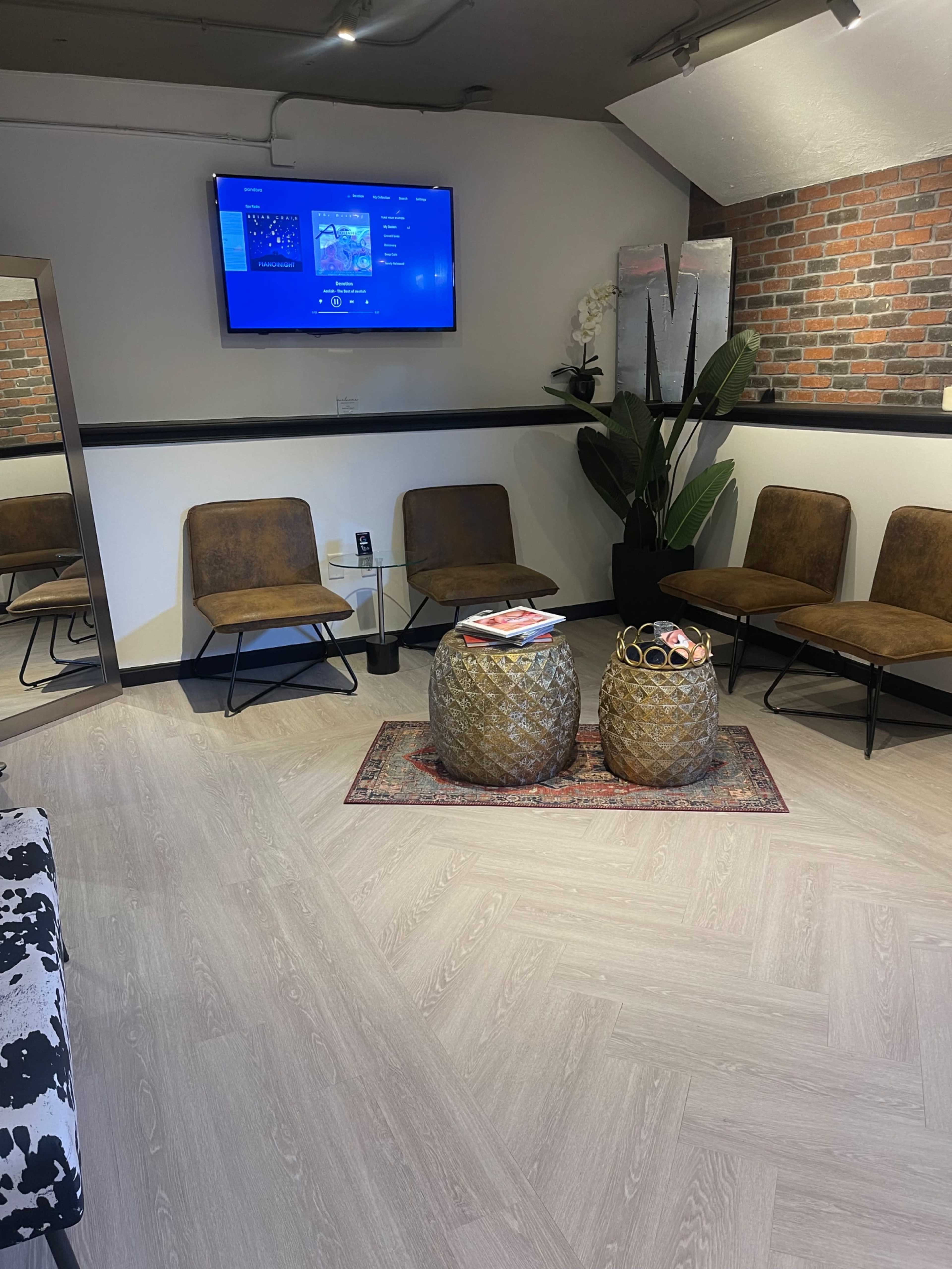 The image shows a waiting area furnished with brown chairs, a small glass table, decorative plant, and a television mounted on the wall, all against a backdrop of brick and wood flooring.