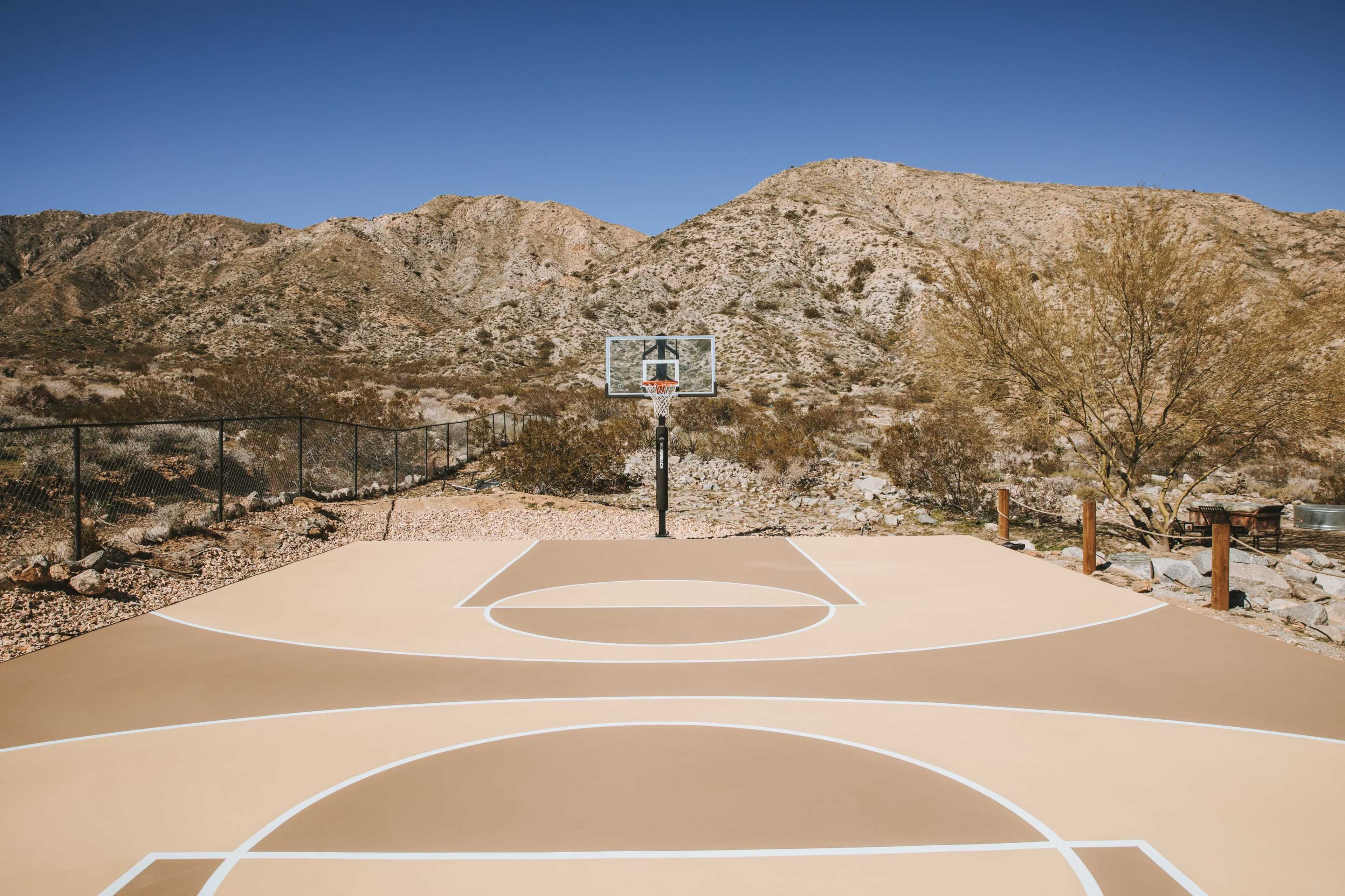 A basketball court with a hoop stands in a desert landscape framed by mountains under a clear blue sky.