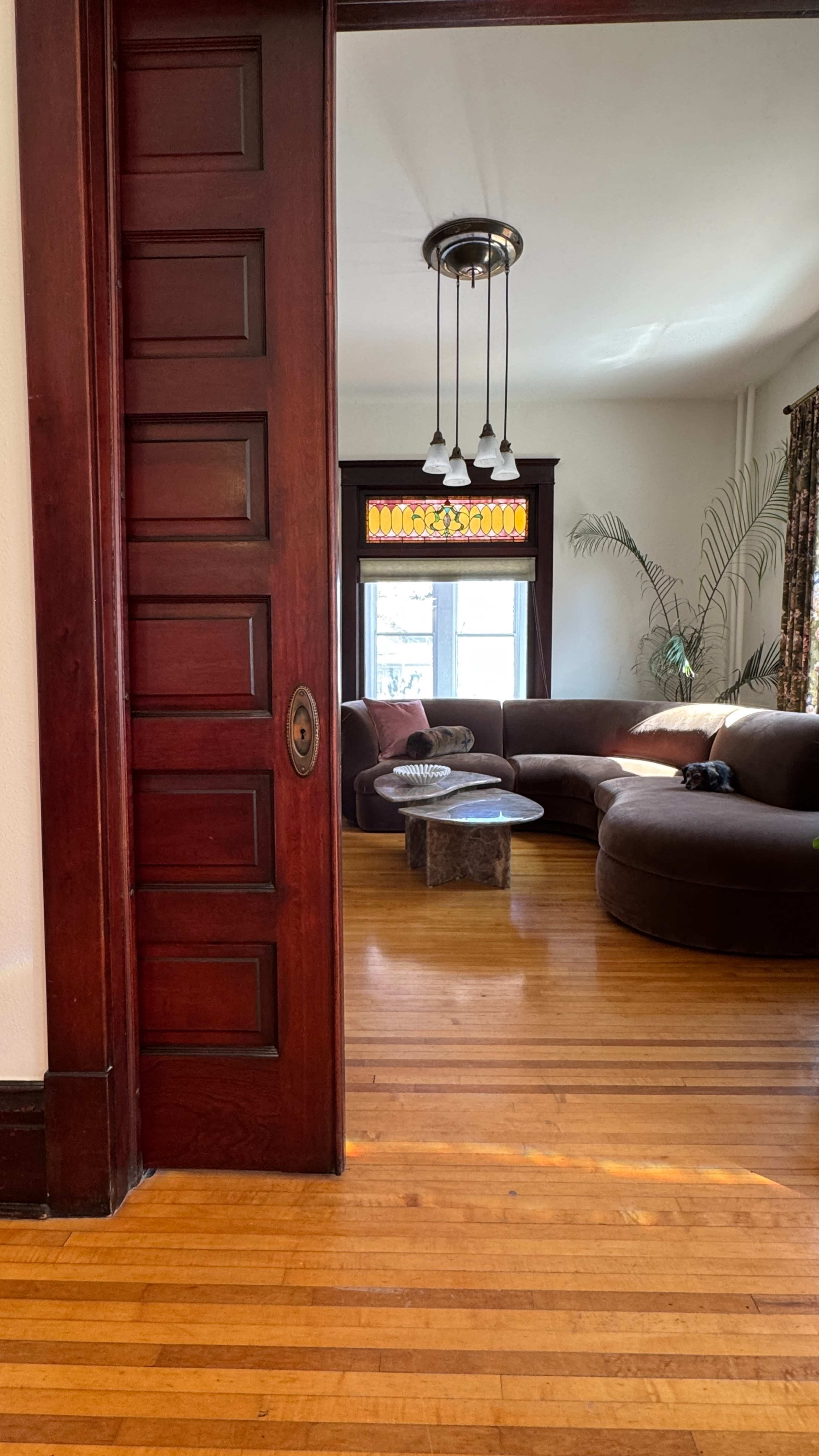 The image shows an interior view of a room with a wooden door, a rounded couch, a coffee table, and a large window with colorful glass above it.